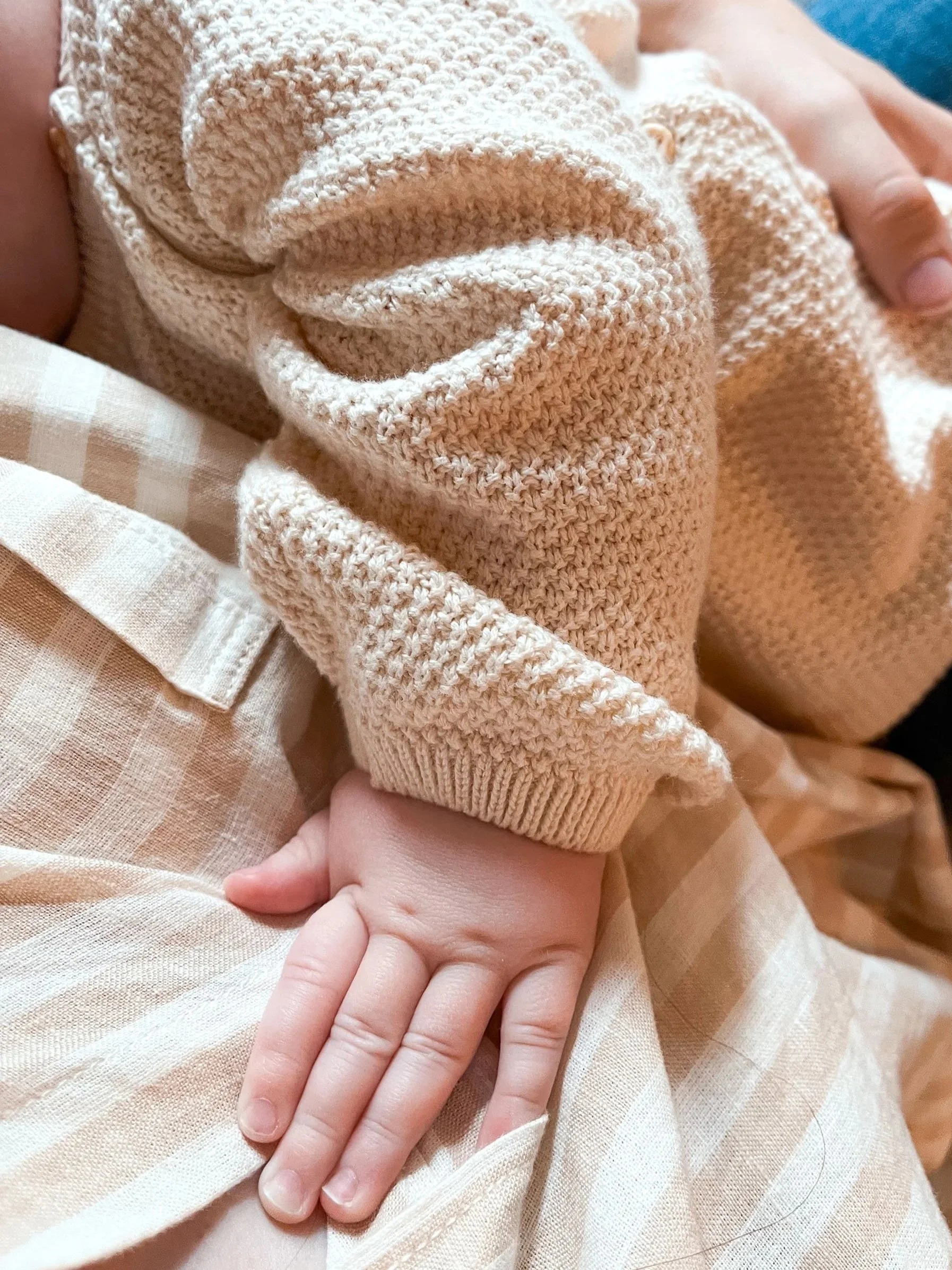 Close-up of a baby's hand resting on a person's lap, with the baby wearing a beige knitted sweater and the person wearing a beige checkered shirt.