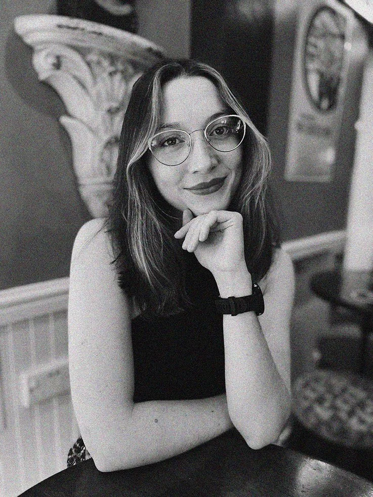 A woman with glasses, shoulder-length hair, and lipstick, sitting at a table with her chin resting on her hand, smiling at the camera.
