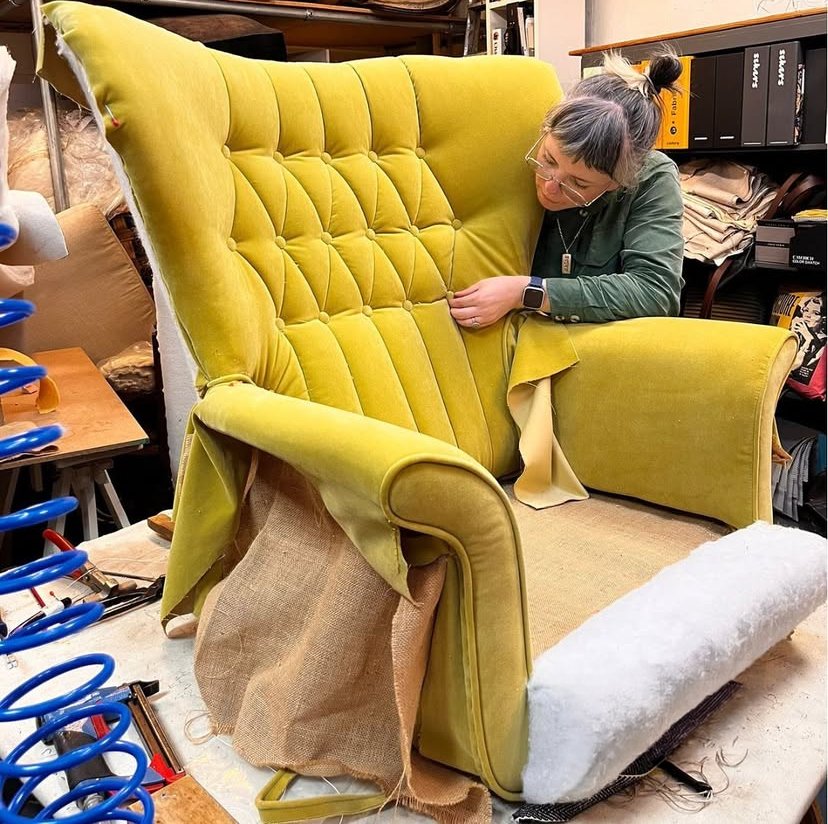A woman in glasses and a green jacket inspects a vintage yellow velvet armchair in a workshop, which is being reupholstered with padding, fabric, and foam.