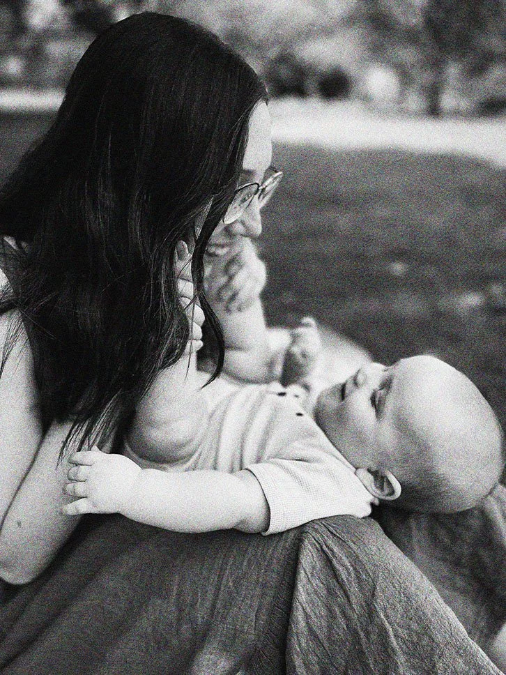 A woman with dark hair and glasses is leaning close to a giggling baby lying on her lap outdoors.