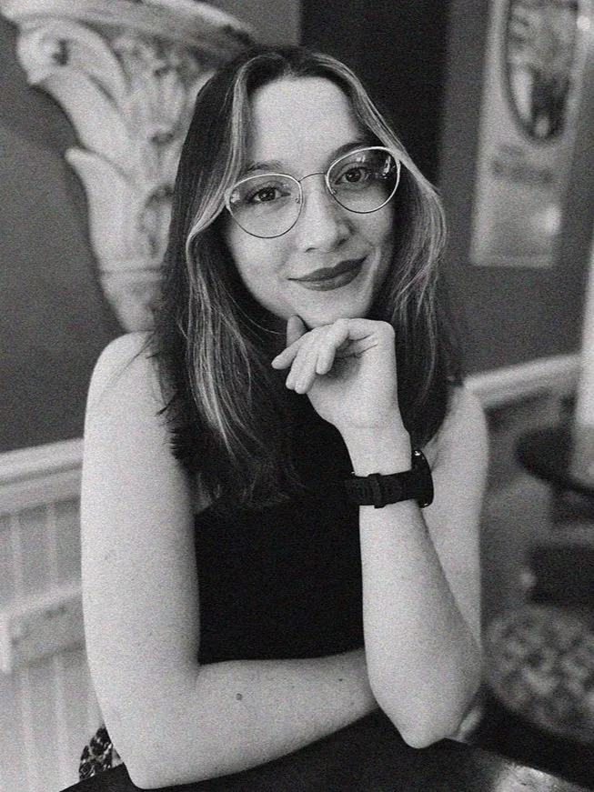 A black and white photo of a woman wearing glasses, smiling, with her chin resting on her hand. She is seated indoors, with a decorative wall and artwork in the background.