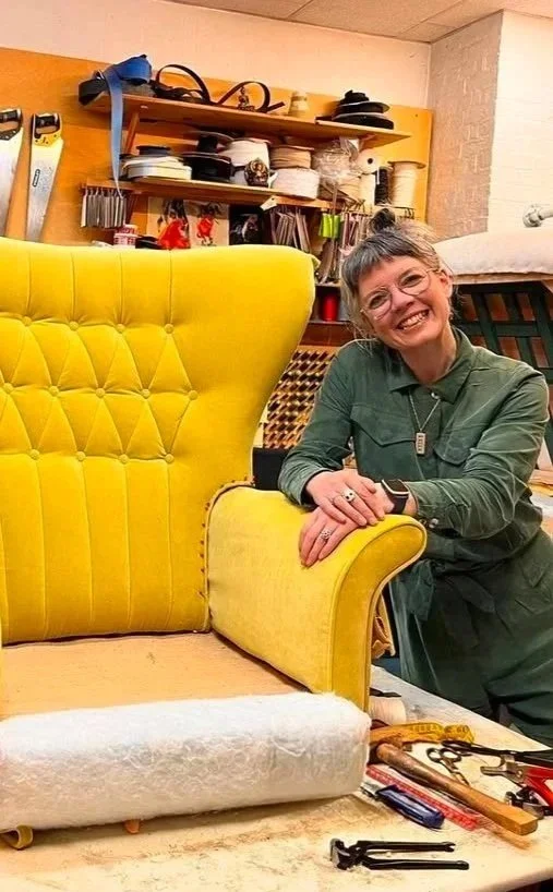 A woman with short gray hair, glasses, and a green shirt is smiling and leaning on a bright yellow, vintage-style upholstered armchair in a workshop with tools, fabric, and shelves in the background.