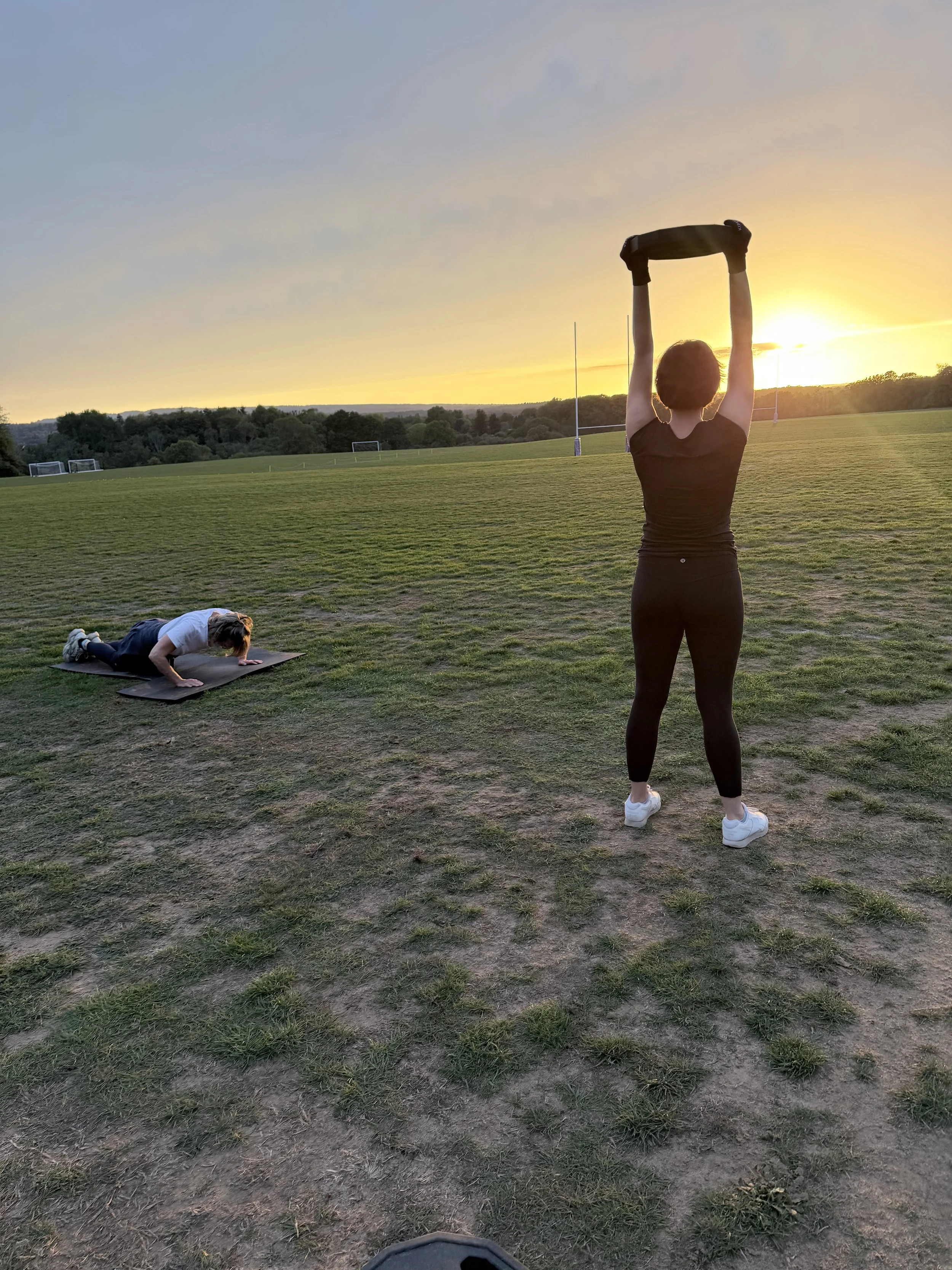 Person standing on a grassy field during sunset, stretching with arms raised overhead while another person is doing push-ups on a mat nearby.