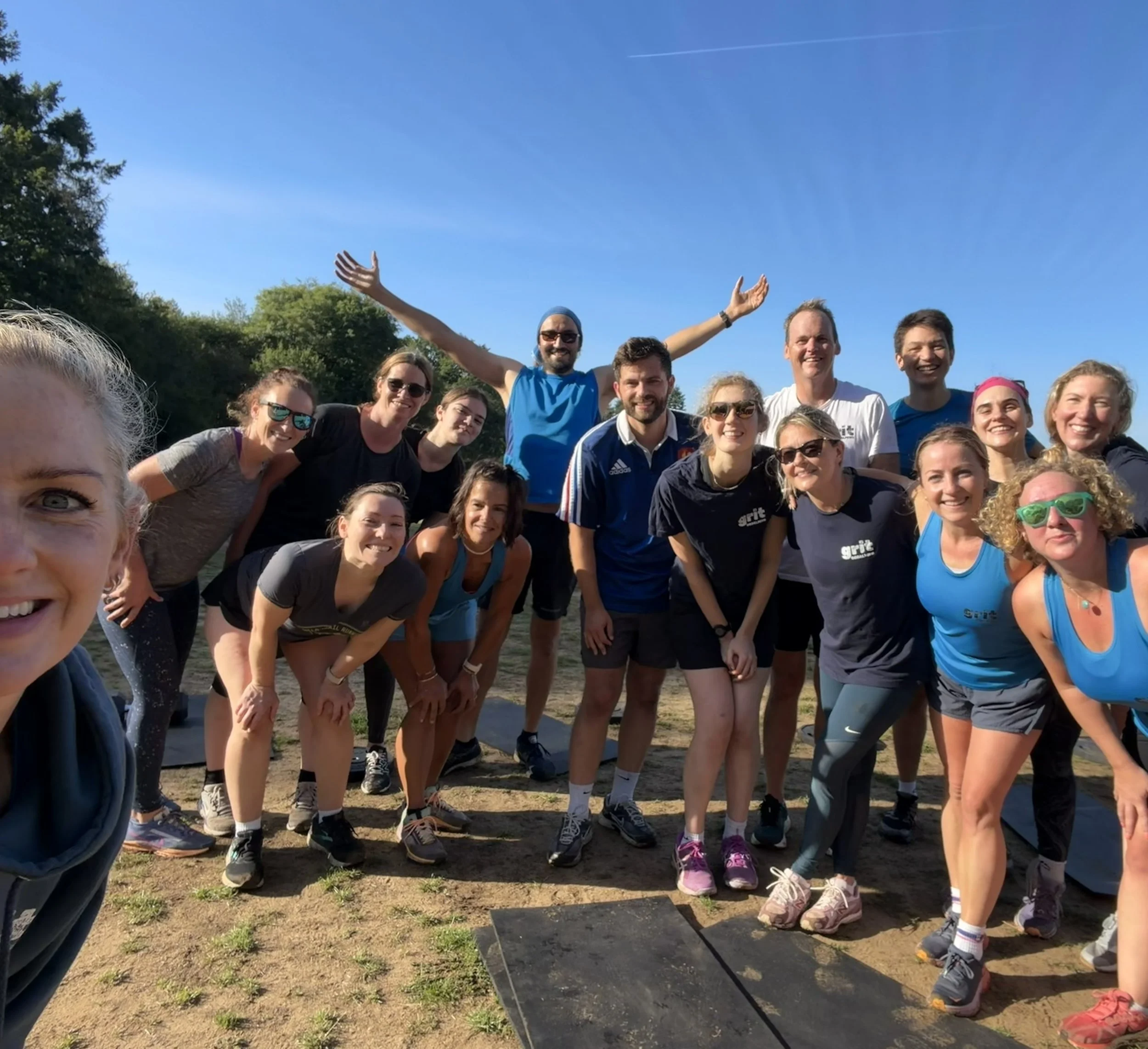 A group of people gathered outdoors on a sunny day, posing together for a photo. Some are smiling and wearing athletic clothes, indicating they might have participated in a running or outdoor event.