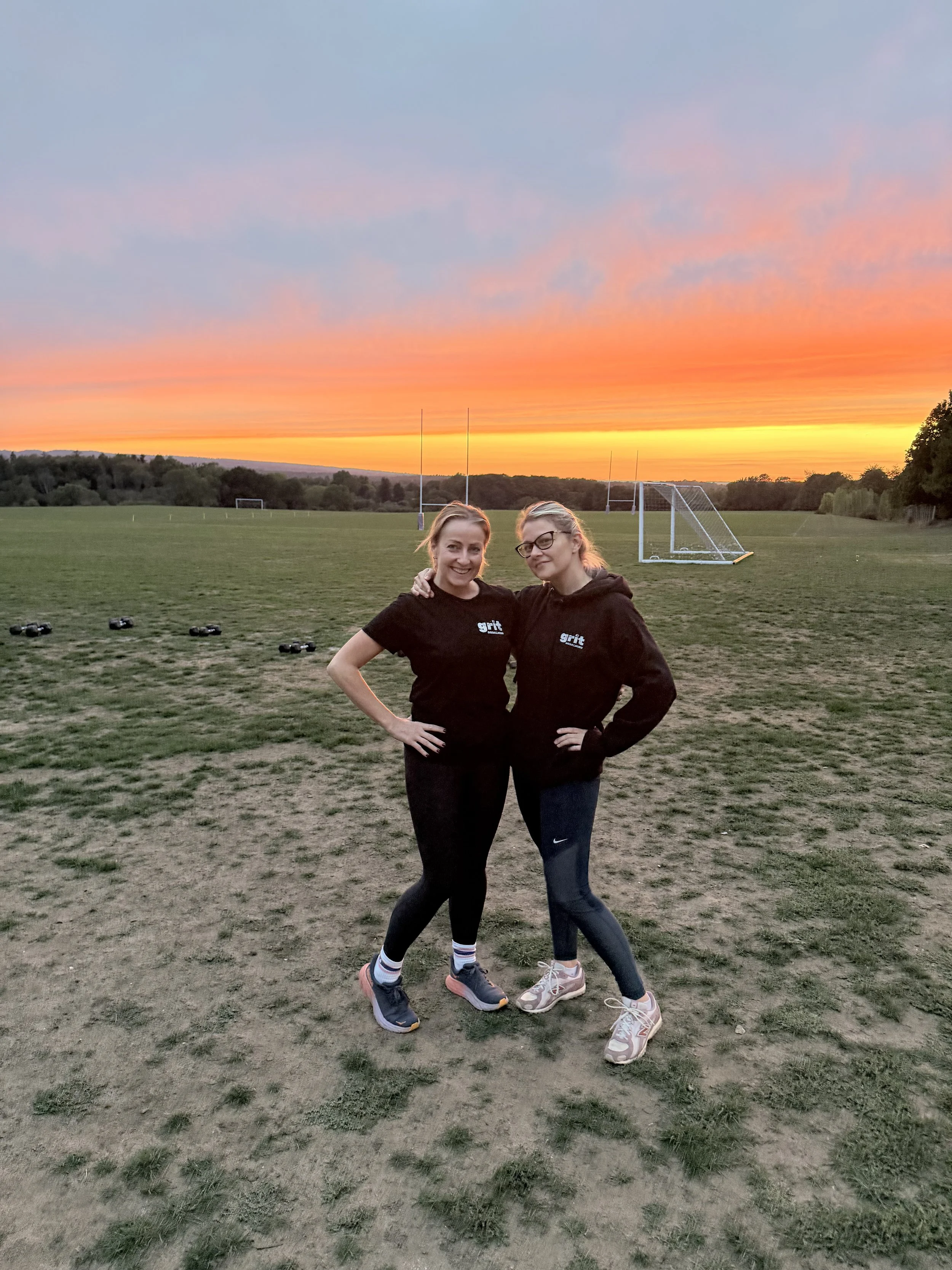 Two women standing together outdoors on a field at sunset, smiling and posing with arms around each other, wearing athletic clothing and trainers, with goalposts and a football placed on the ground behind them.