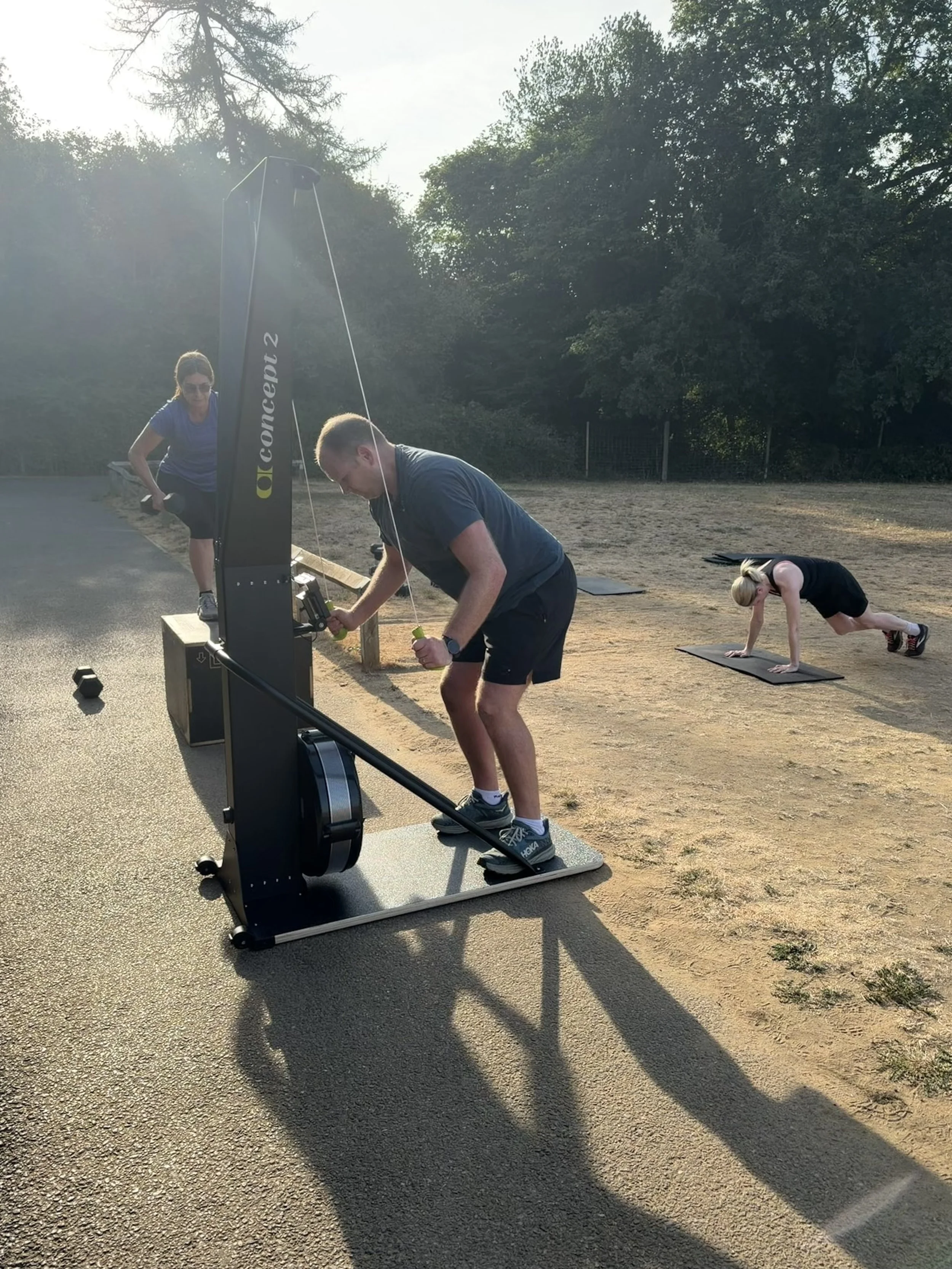 People exercising outdoors, with one person using a rowing machine and others doing floor exercises on mats, in a park area with trees in the background during late afternoon or early evening.