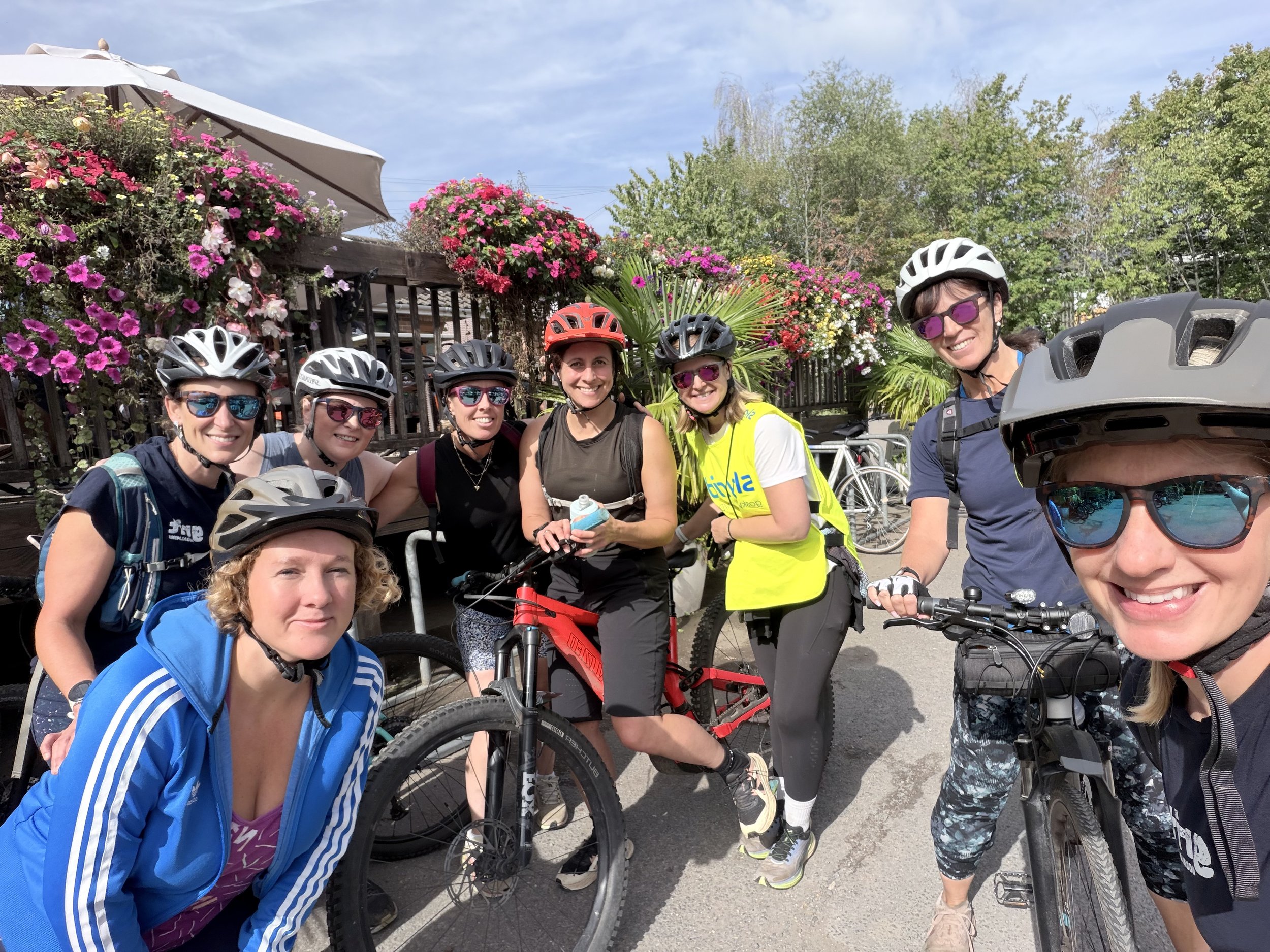 A group of seven women wearing cycling helmets and sunglasses, standing with their bikes in front of a fence with colorful flowers and green trees, under a partly cloudy sky.