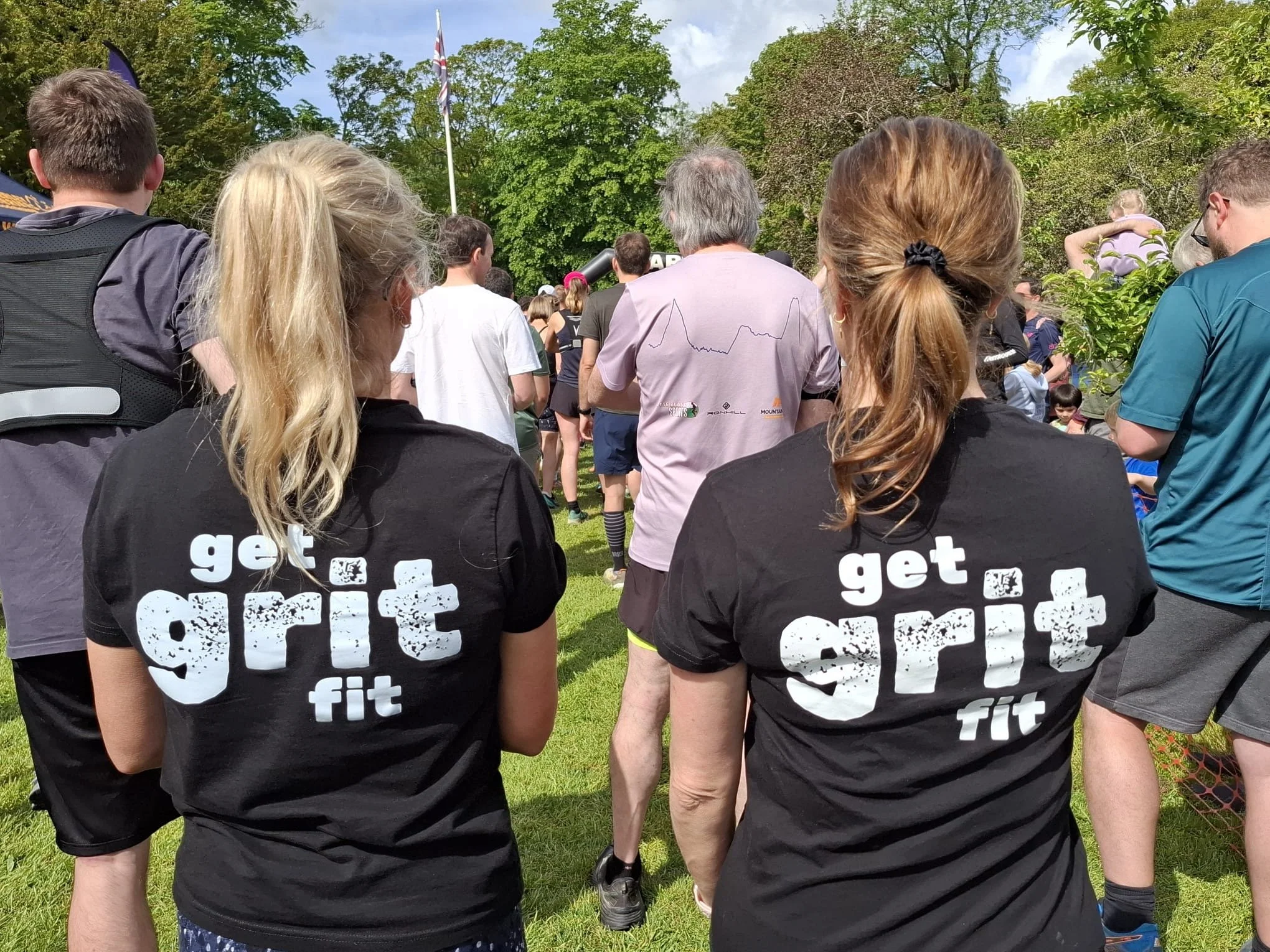 Two women with branded black t-shirts standing side by side, with the shirts reading 'get grit fit' in white text. They are outdoors among a crowd of people, with trees and a clear sky in the background.