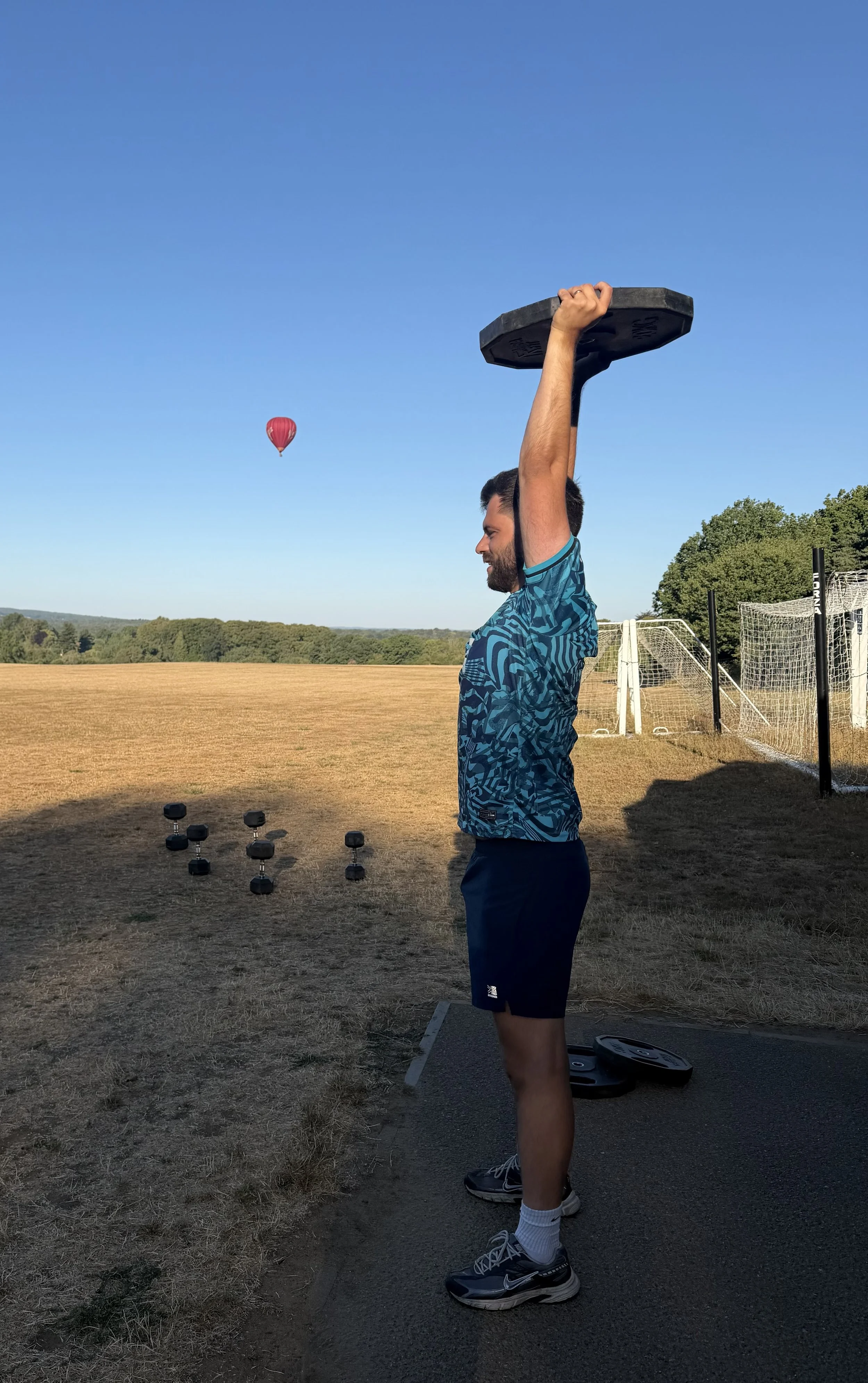 A man in a blue t-shirt and black shorts lifts a weight plate overhead outdoors during sunset. In the background,  on Godalming College sports field with football goals, a group of small dumbbells on the ground, and a hot air balloon in the sky.