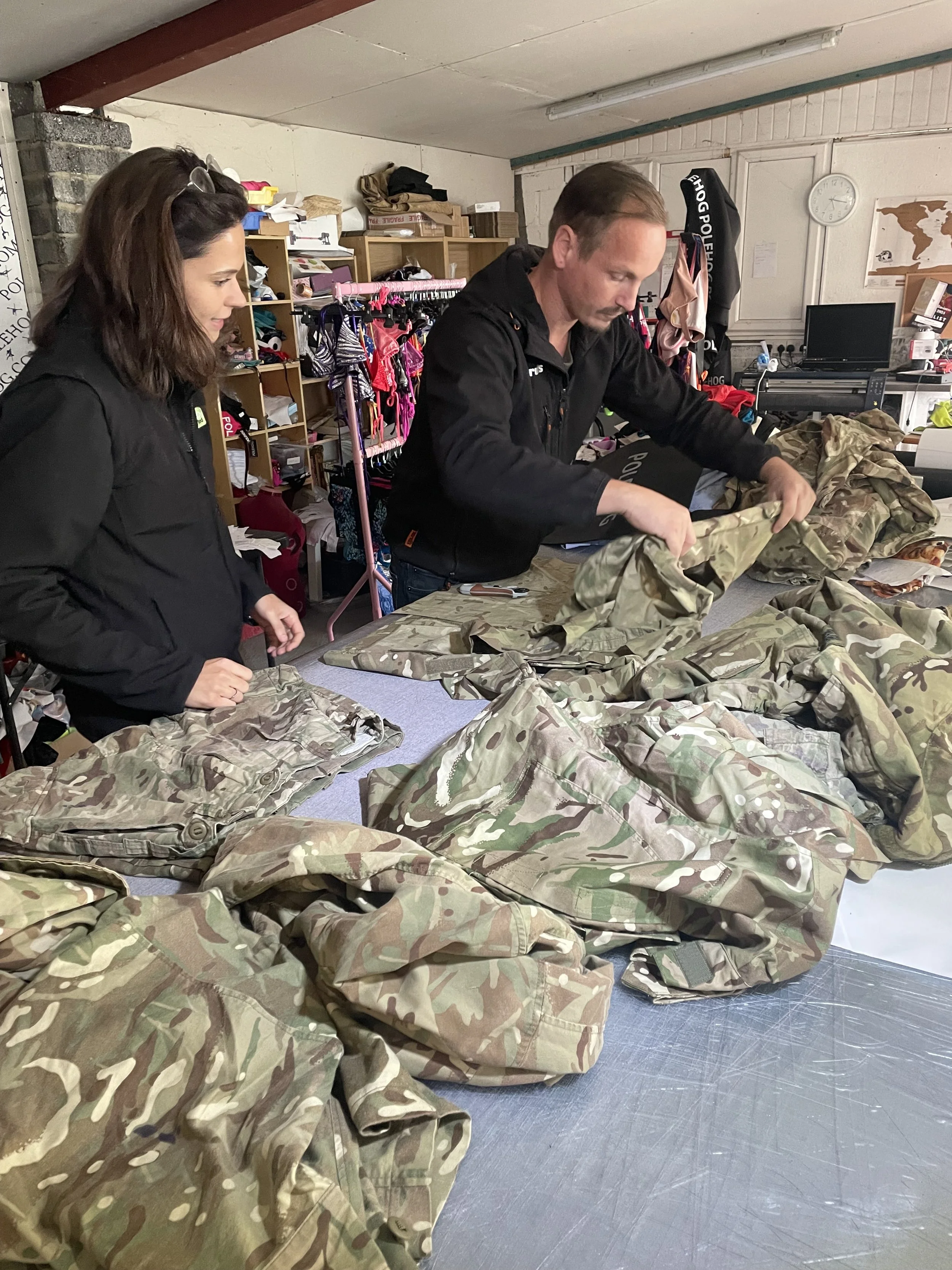 Two people inspecting camouflage military uniforms on a table in a warehouse or clothing store.
