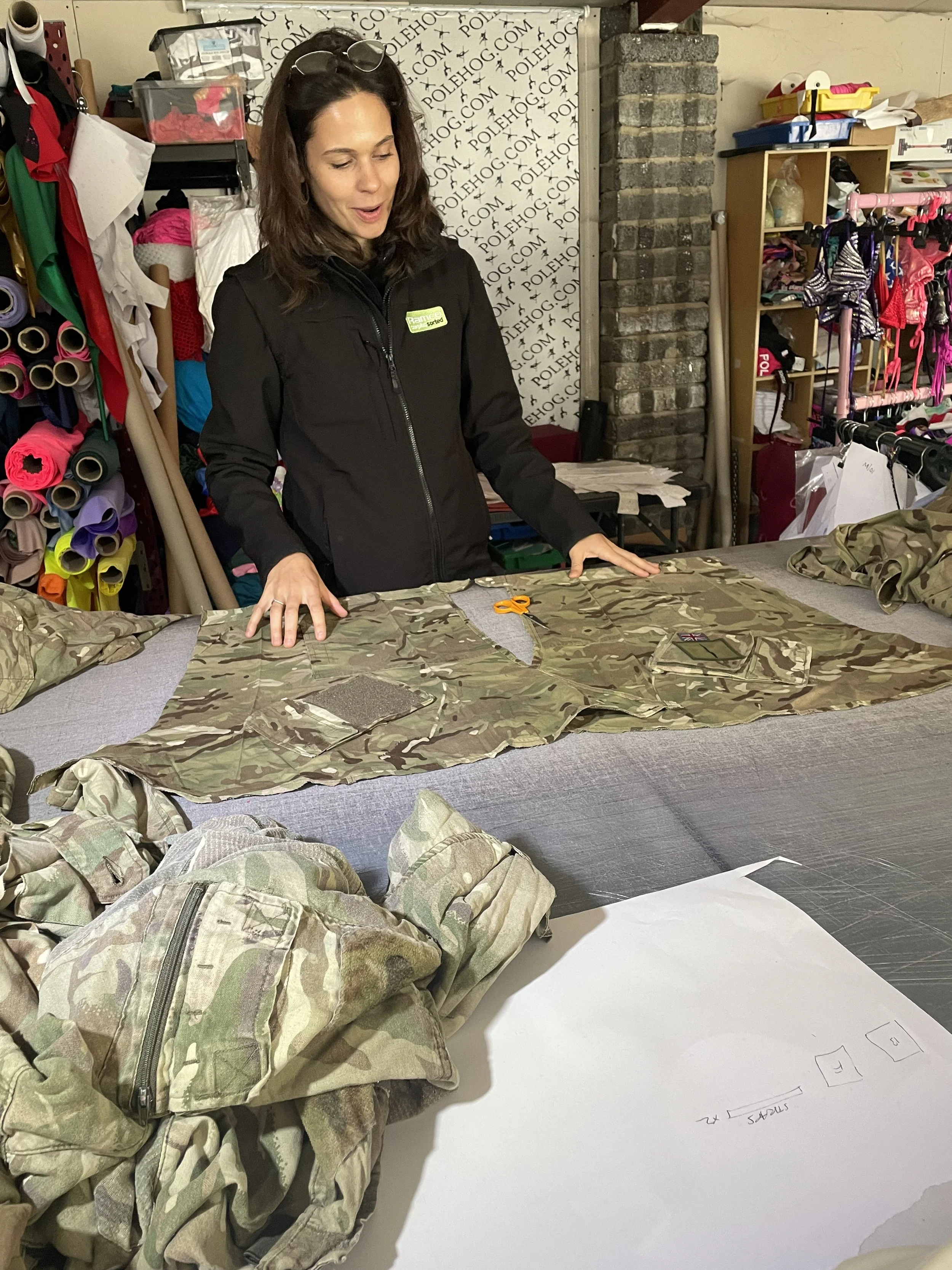 A woman in a black jacket is standing at a table with camouflage fabric and clothing, in a workshop or tailoring space with shelves filled with fabric, clothing, and accessories in the background.