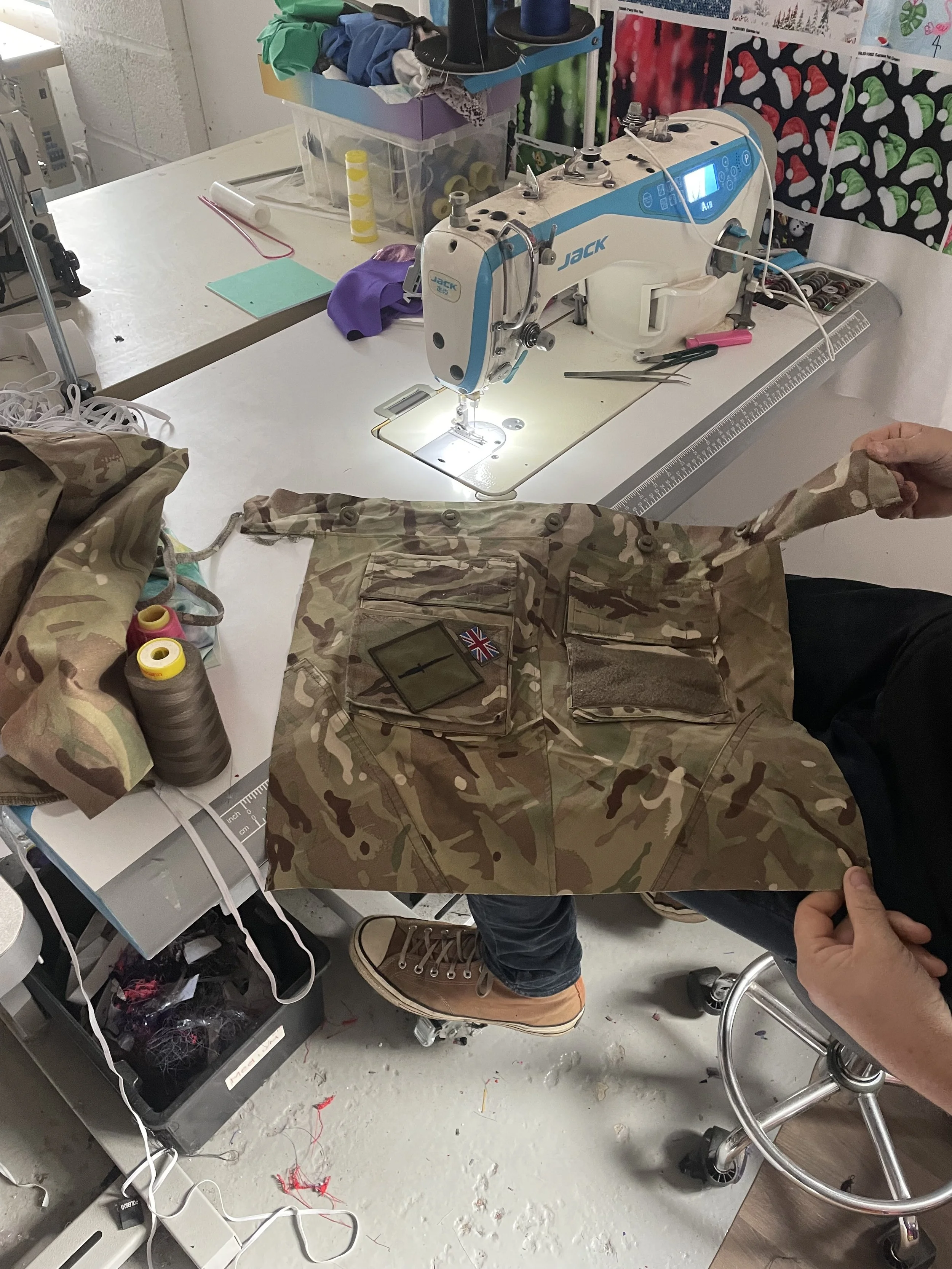 A person holding up a camouflage military uniform in front of a sewing machine in a workshop. The sewing station has various supplies including threads, fabric, and a clear storage box with more sewing materials.