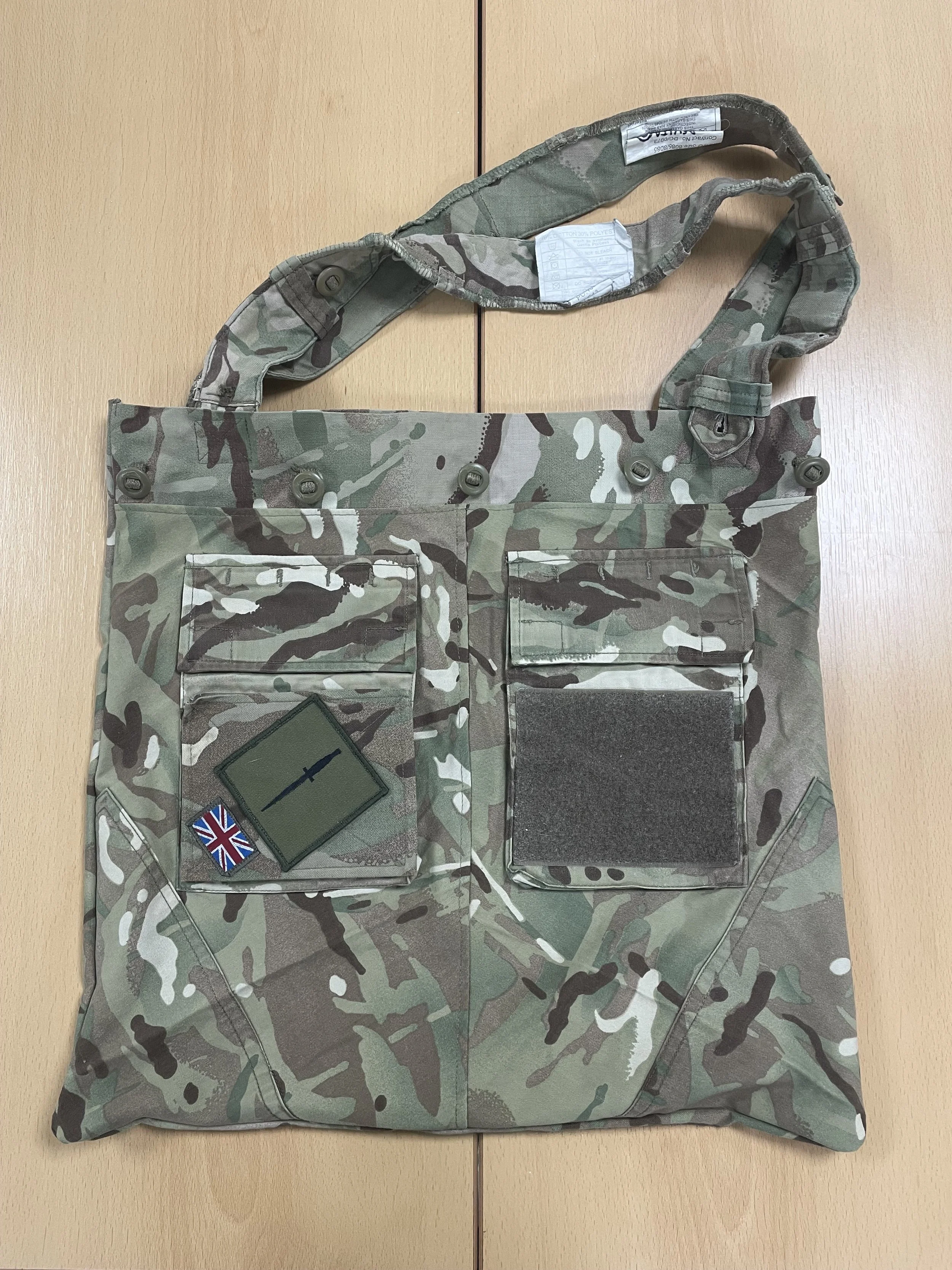 Camouflage tactical bag with patches, including a Union Jack flag patch and a black sword insignia patch, laid flat on a wooden surface.