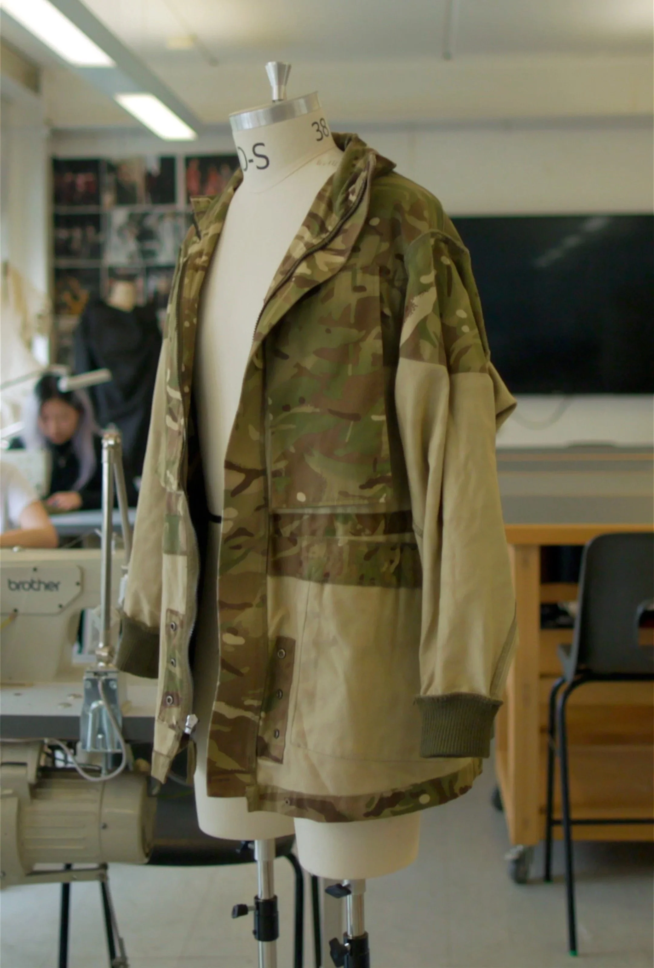 A mannequin wearing a camouflage jacket and a beige skirt in a classroom or workshop setting with tables, chairs, and people in the background.