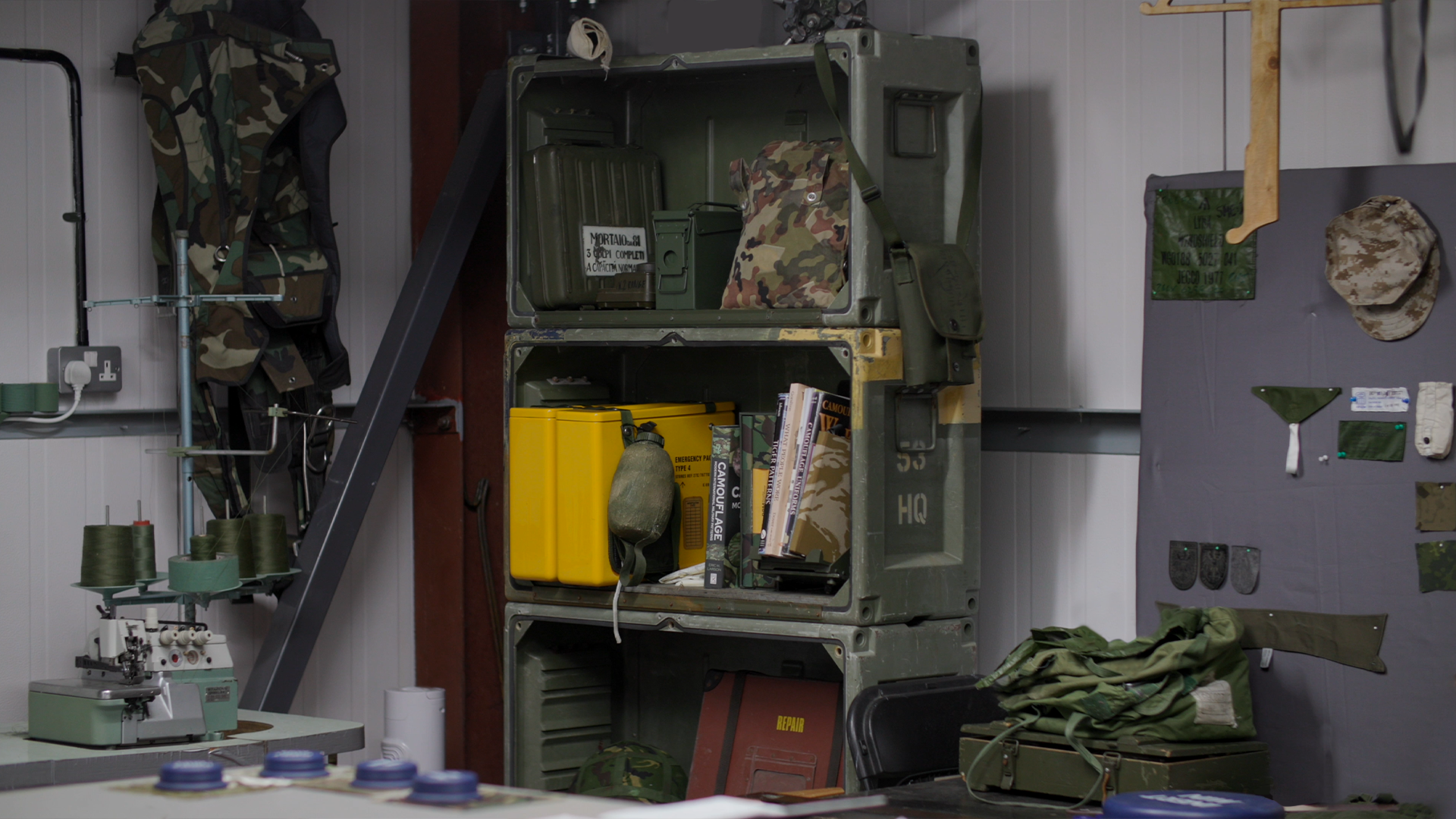 A military storage room with shelves holding equipment, including camouflage gear, a yellow case, and books. The room has wall-mounted patches and gear, with a sewing machine and spools on a table in the foreground.