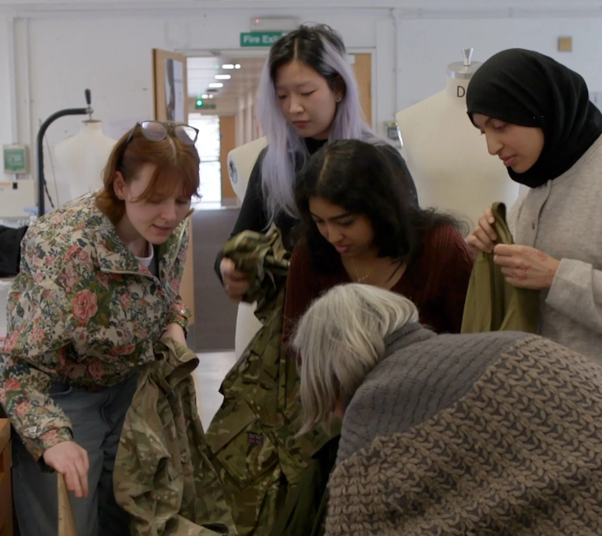 Group of women gathered around a table, examining something, in a room with medical or laboratory equipment, including a mannequin head and a fire exit sign.