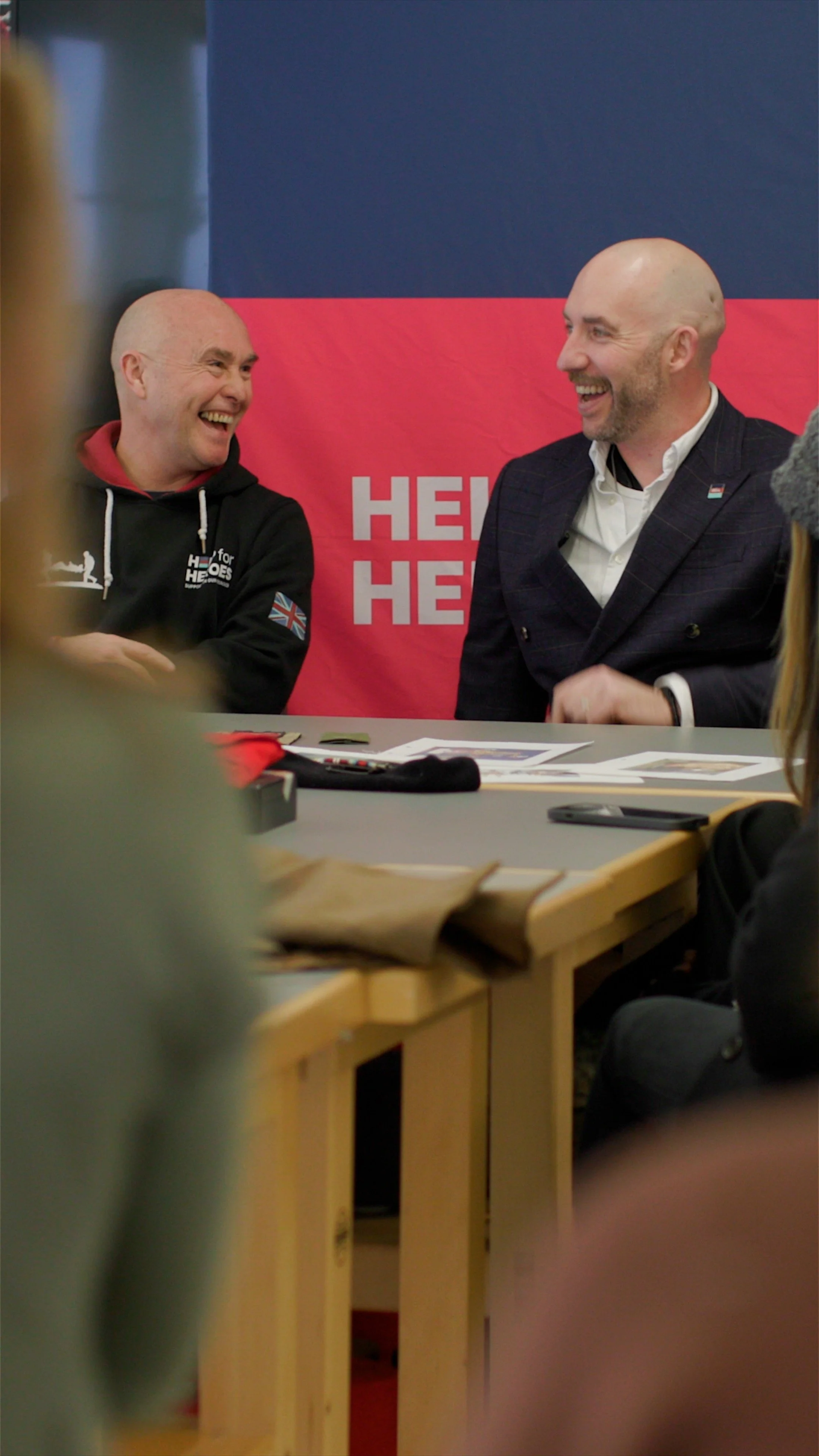 Two men sitting at a table, smiling and talking, with a red and blue background behind them.