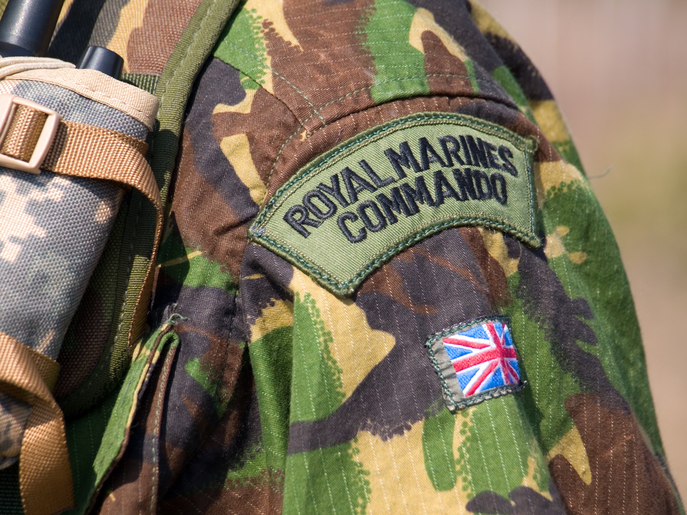 Close-up of a British DPM camouflage uniform with a "Royal Marines Commando" shoulder patch and Union Jack tab, highlighting the iconic green, brown, black, and tan Disruptive Pattern Material used by the UK Armed Forces.