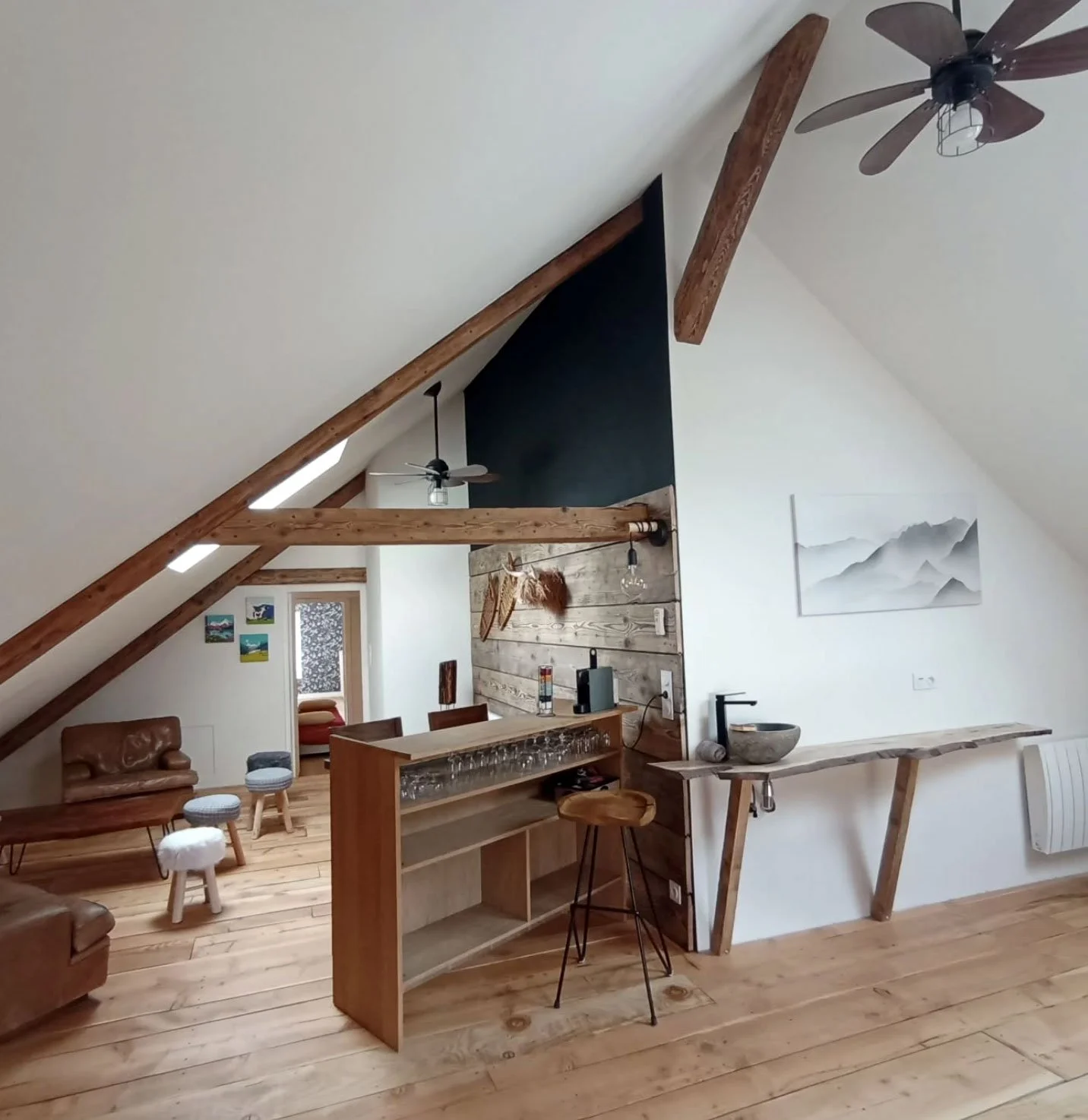 Intérieur d'un loft avec plafond en pente, mobilier en bois, canapés en cuir, bar avec des verres, décor rustique et moderne, avec des ventilateurs de plafond, une œuvre d'art de montagne, et un sol en bois.