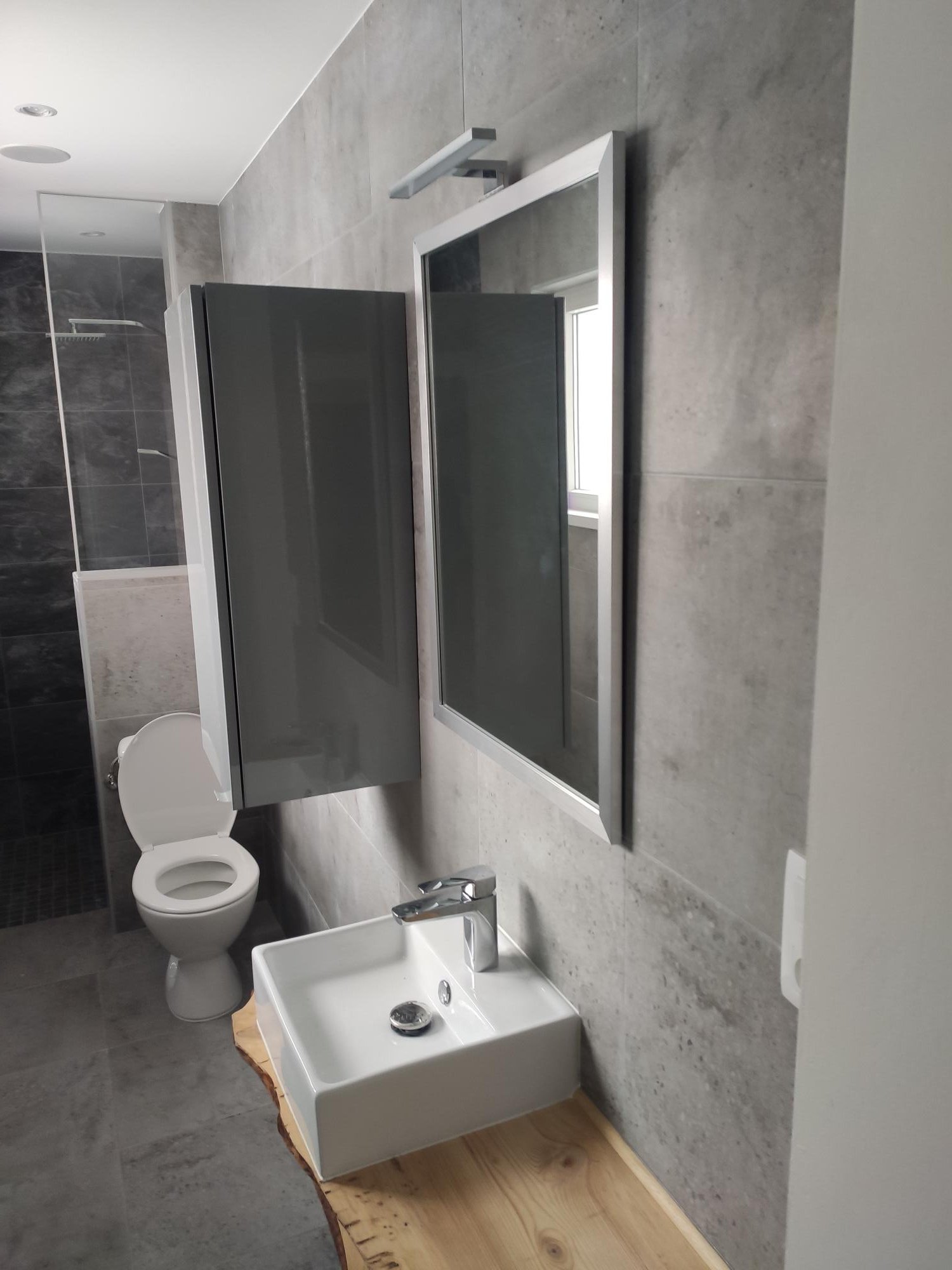 Petit lavabo blanc avec robinet, miroir, toilette avec couvercle relevé, armoire grise au mur, murs en carrelage gris, cabine de douche avec paroi en verre dans une salle de bain moderne.