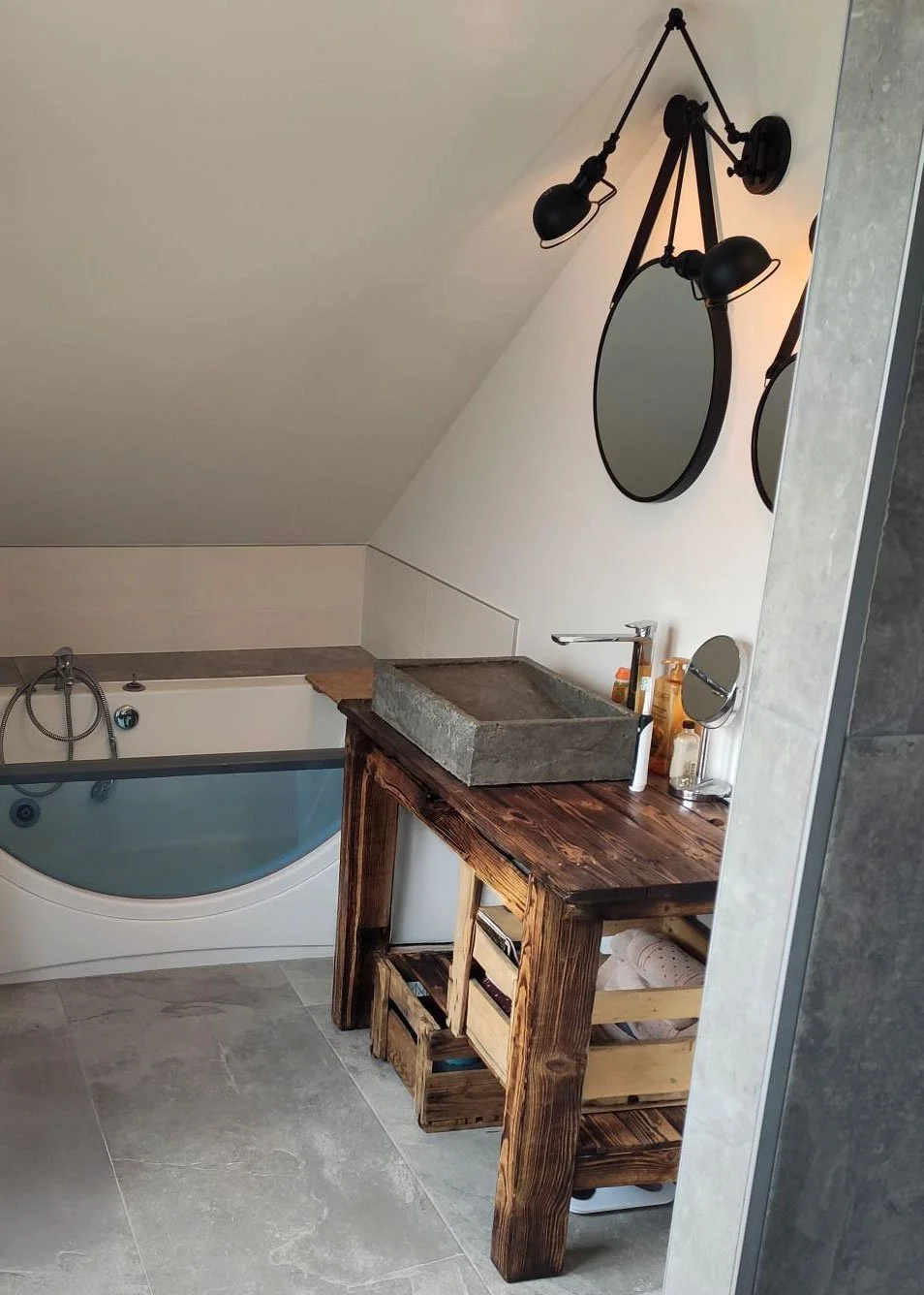 Une salle de bain avec une baignoire, un lavabo carré en pierre sur un meuble en bois rustique, deux miroirs ronds, deux lampes murales noires et quelques produits de toilette sur le comptoir.