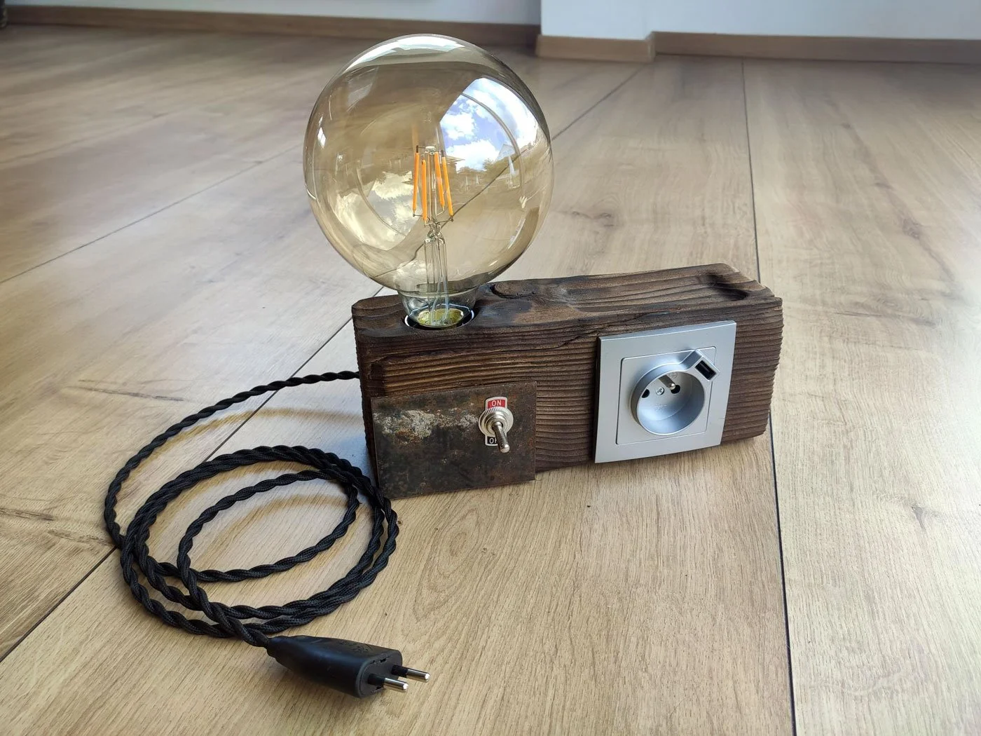 Ampoule électrique ancienne fixée sur une planche en bois, avec un interrupteur et une prise électrique, posé sur un sol en bois.