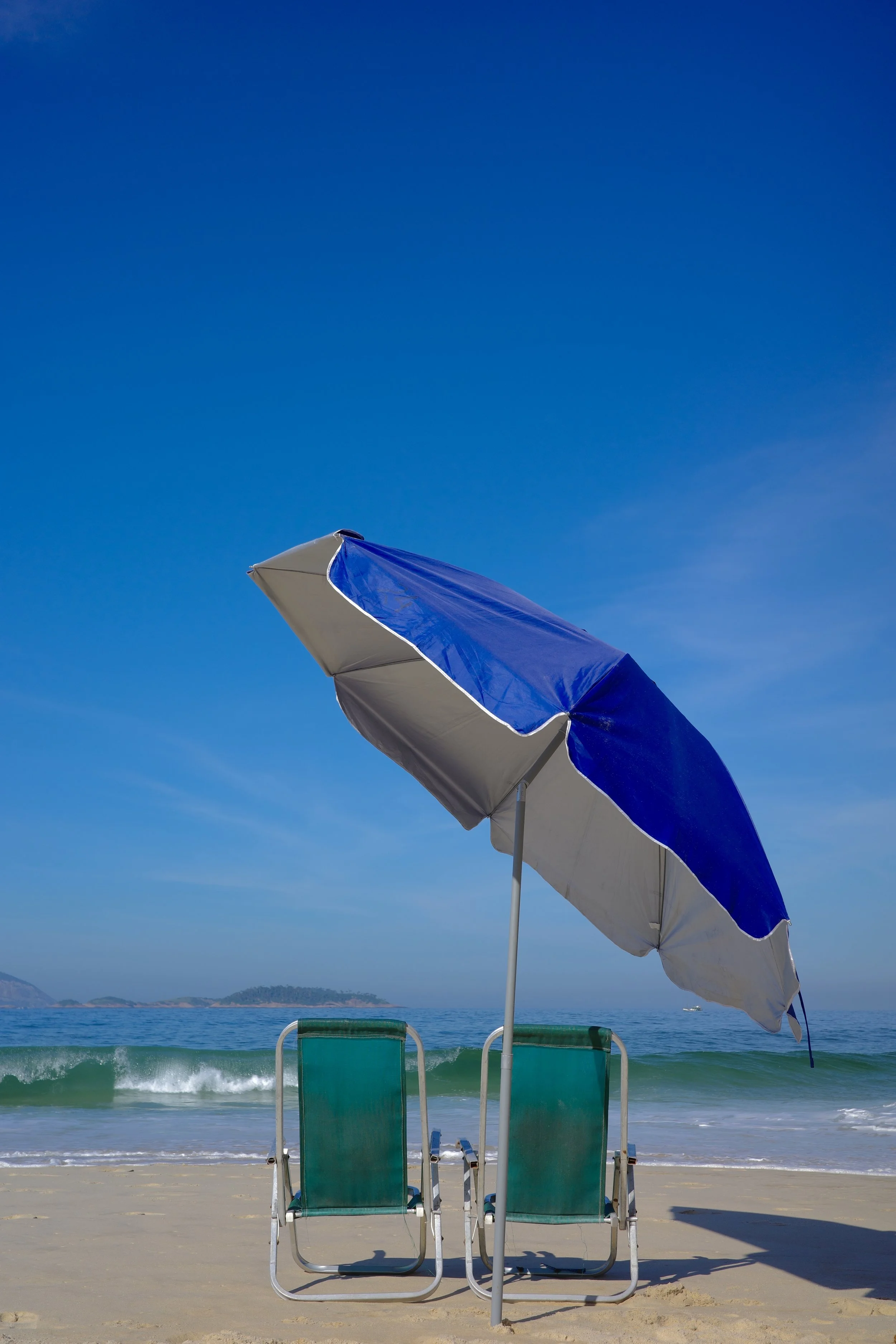 Paréo bleu et blanc, parasol rayé bleu et blanc, deux chaises longues sur la plage avec vue sur la mer et des montagnes à l'horizon.