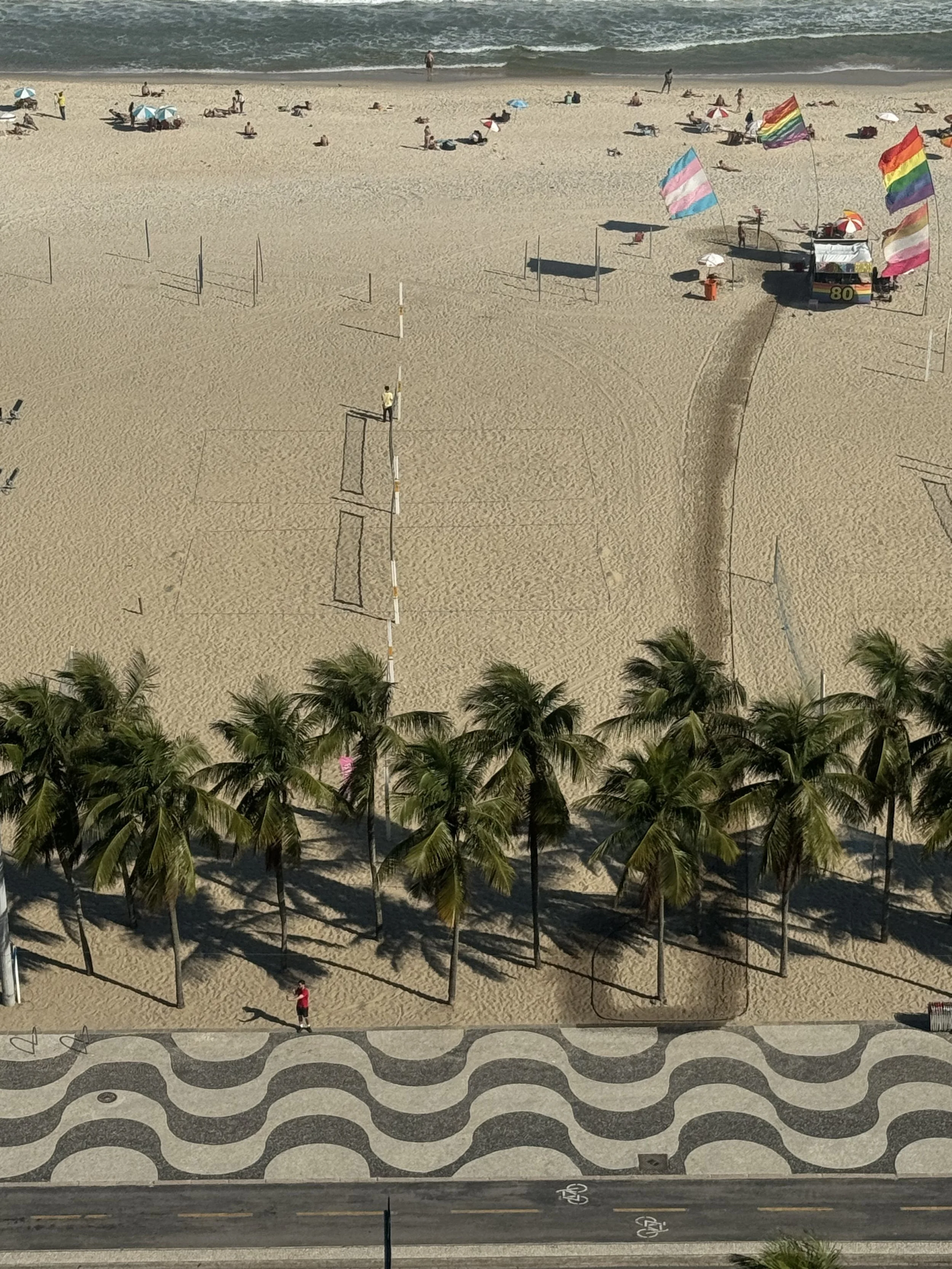 Plage de sable avec des parasols, des gens qui se détendent, un stand de fruits ou de souvenirs avec des drapeaux arc-en-ciel, et une rangée d'arbres en contrebas