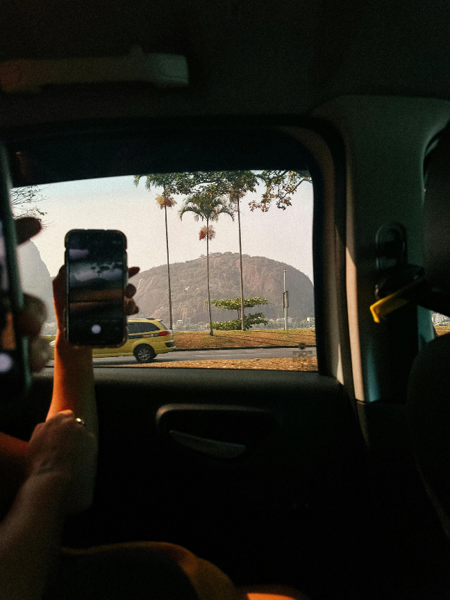 Vue depuis l'intérieur d'une voiture montrant un paysage avec une montagne, des palmiers et une voiture jaune passant devant. Une personne tient un téléphone pour prendre une photo du paysage.