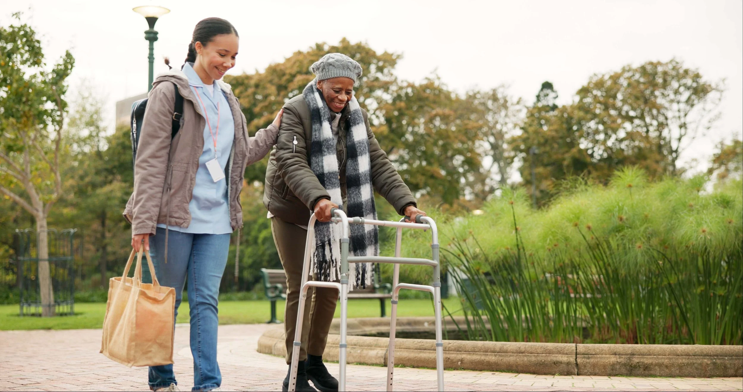 A young woman helping an elderly woman walk with a walker in a park.