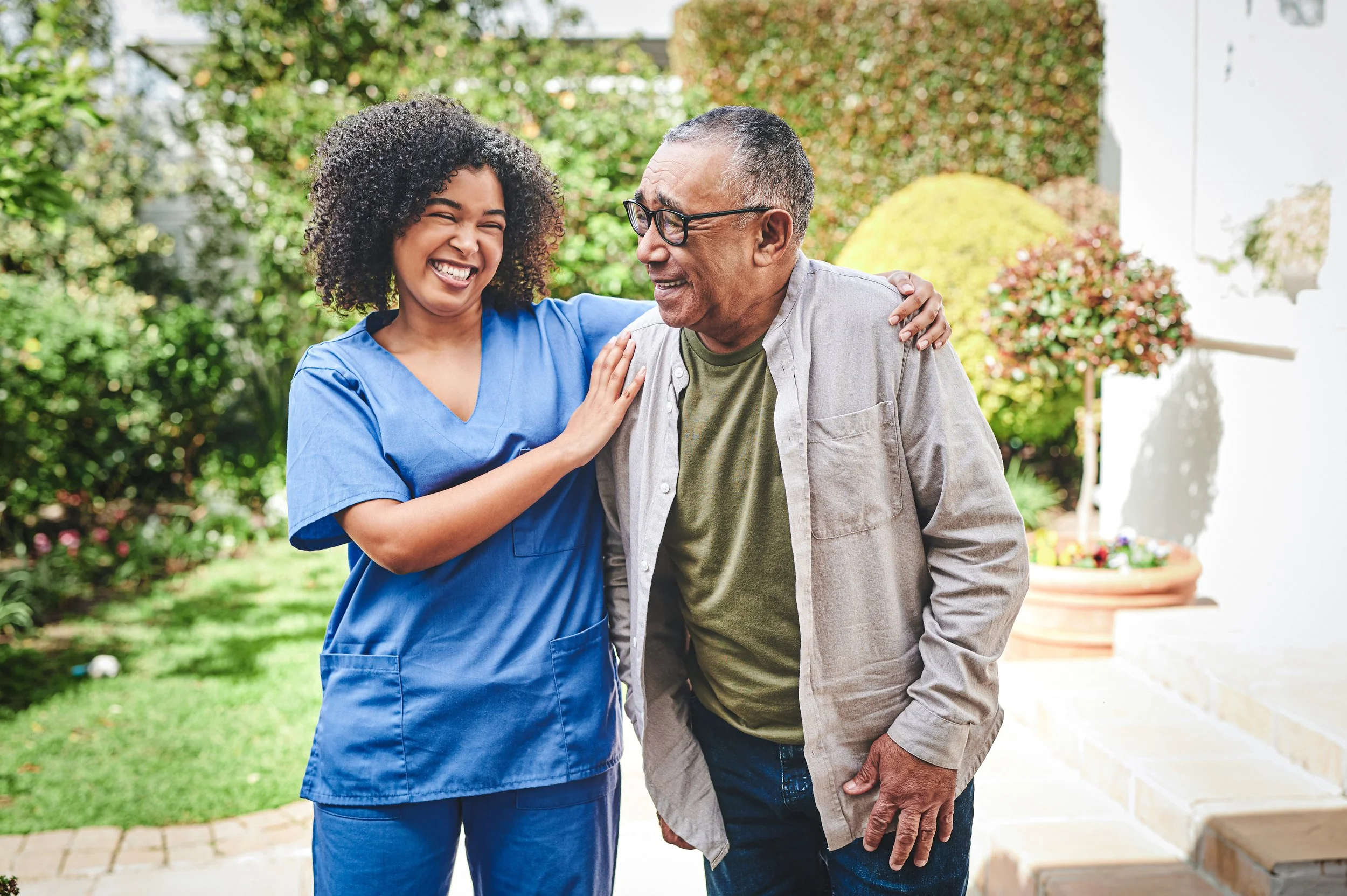 A nurse in blue scrubs smiling and comforting an elderly man with glasses, outdoors in a garden.
