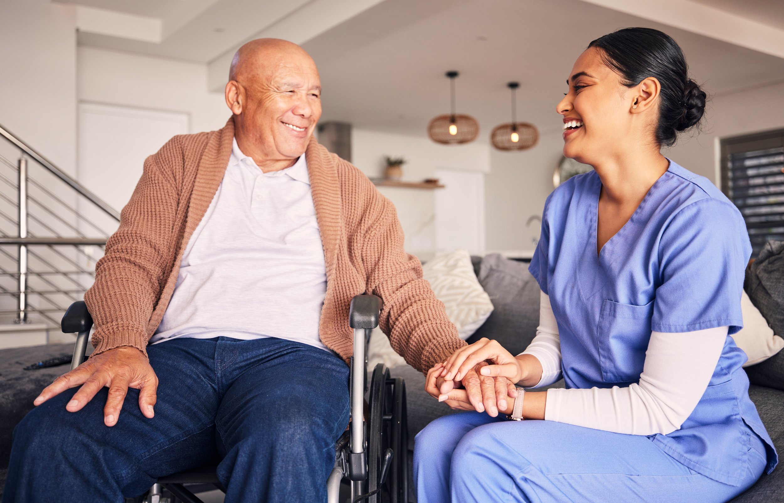 A healthcare worker in blue scrubs holding hands with an elderly man in a wheelchair, both smiling and enjoying a moment together in a cozy living room.