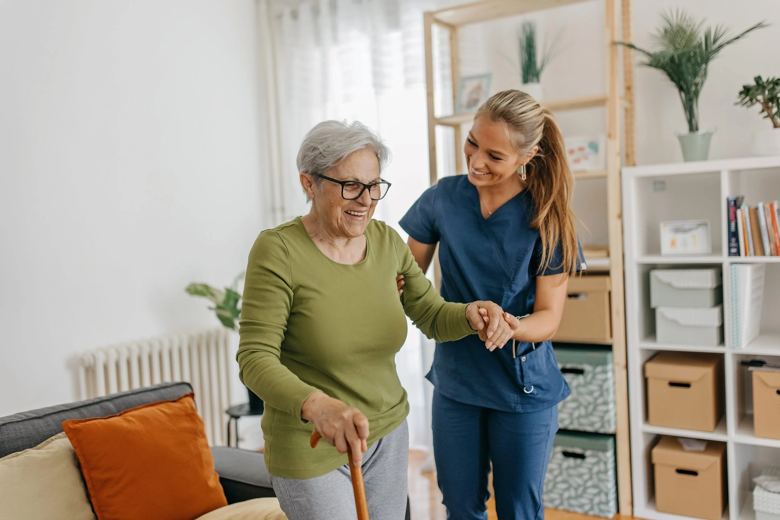 A young nurse assisting an elderly woman with a cane, helping her stand or walk inside a well-lit living room.