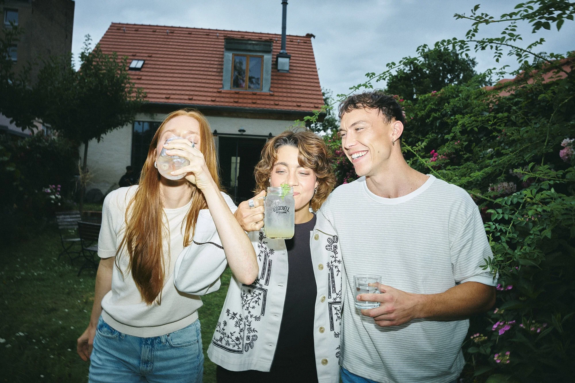 Three friends gather outdoors in the evening, enjoying drinks and smiling together. One woman with long red hair is drinking from a glass, the woman with short brown hair is holding a mug, and the man with short dark hair is holding a clear glass of water, all standing close to each other with a house and greenery in the background.