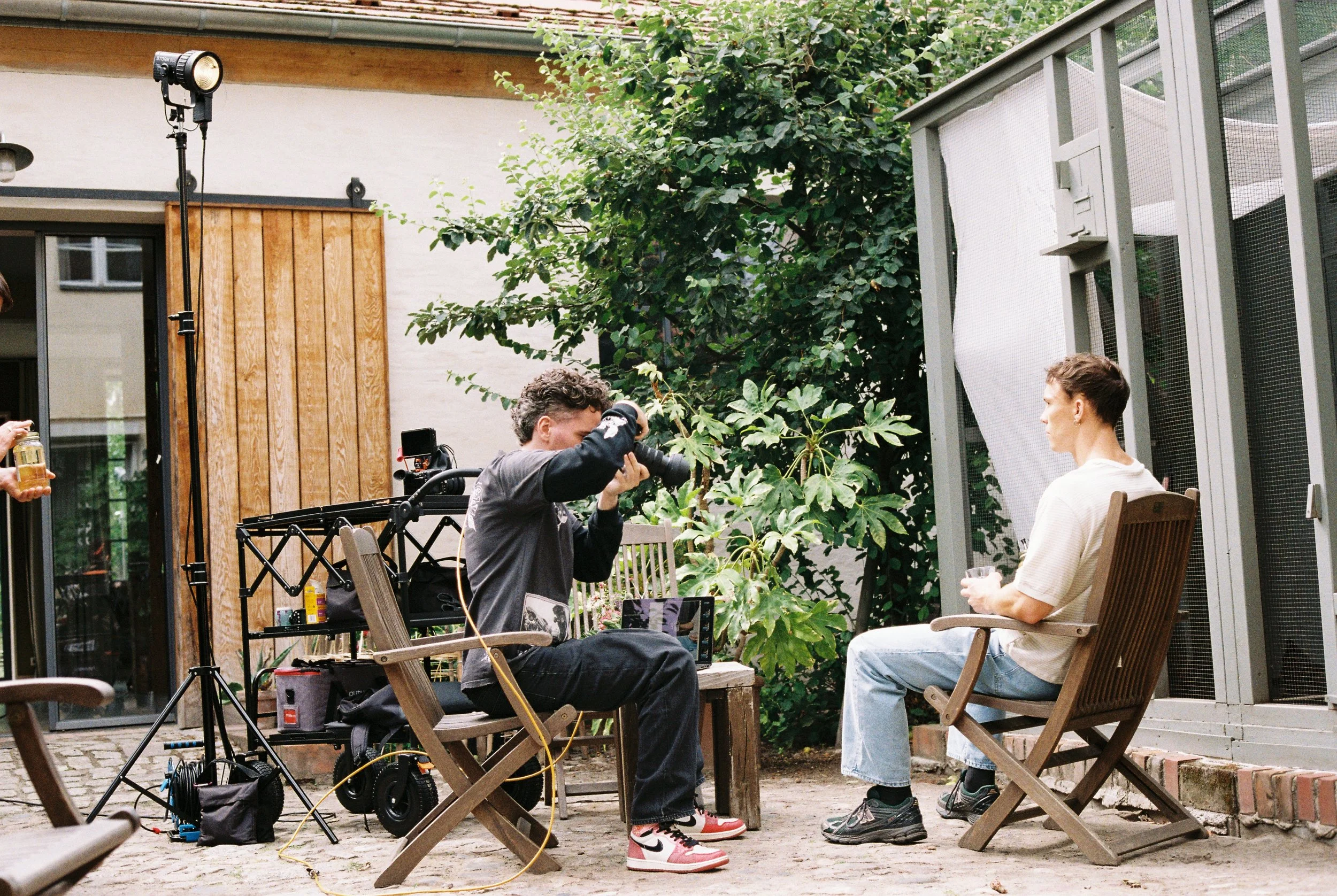 Two people sitting on wooden chairs and having a conversation outdoors in a garden or patio area, surrounded by trees and plants, with film or photography equipment nearby.