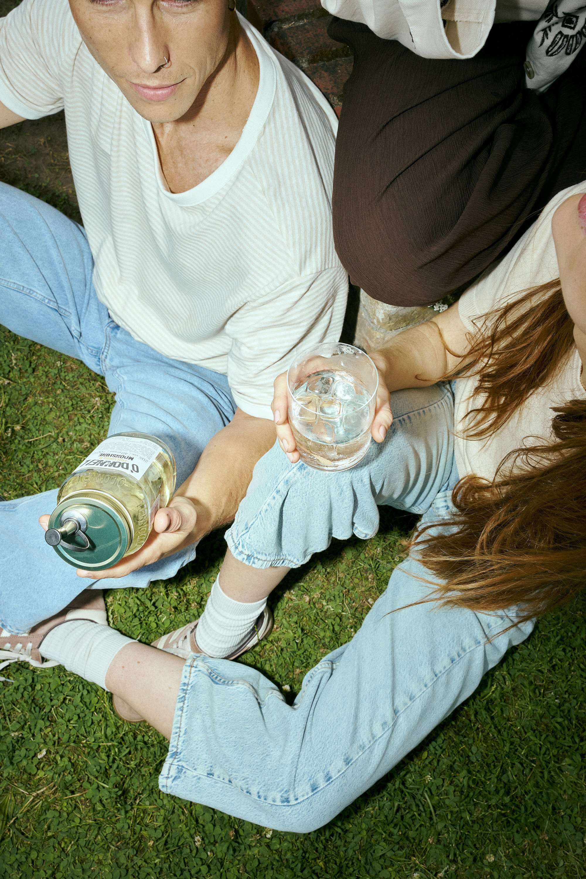 A group of people sitting on grass, partially visible, holding drinks with ice, with one person reaching for a bottle. The individuals are wearing casual clothing including jeans and t-shirts, and the scene has a relaxed and social atmosphere.