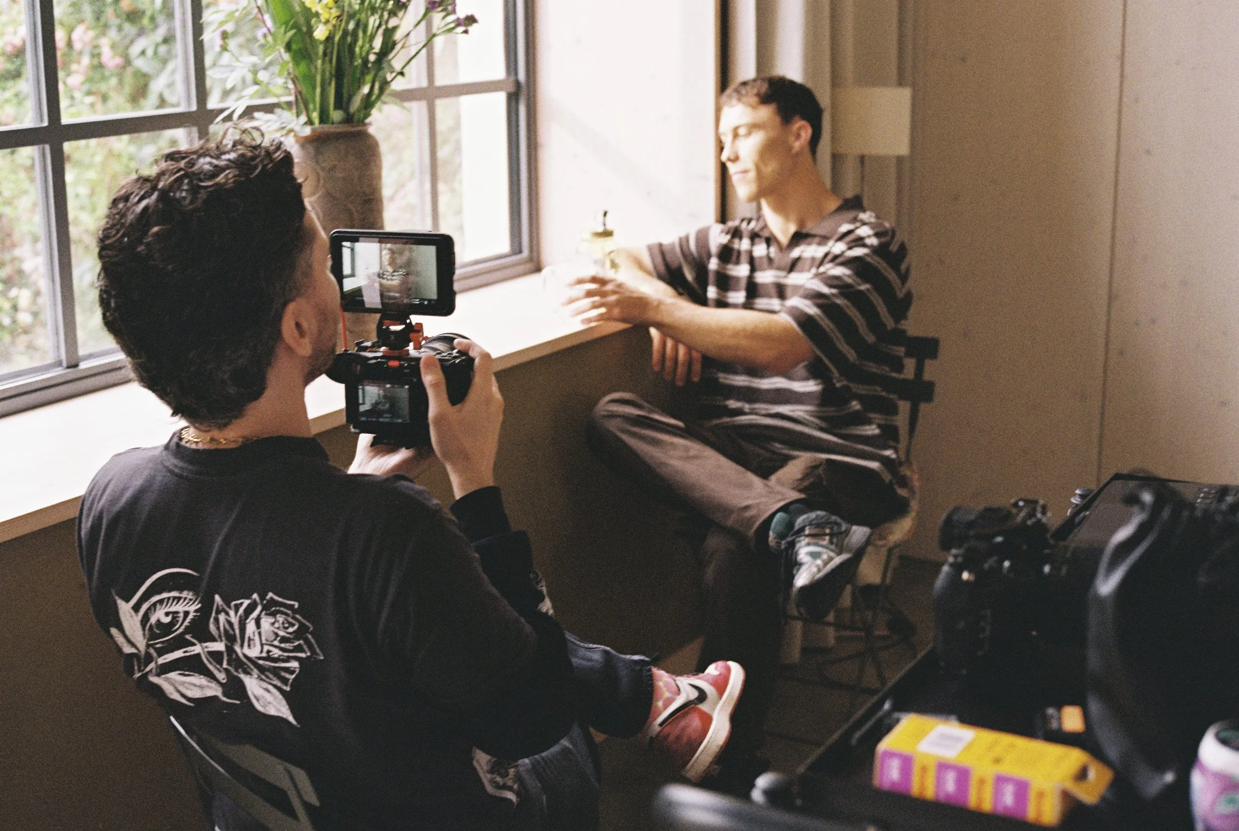 A person filming another person sitting on a window ledge with a camera mounted on a stabilizer, in a room with large windows and natural light.