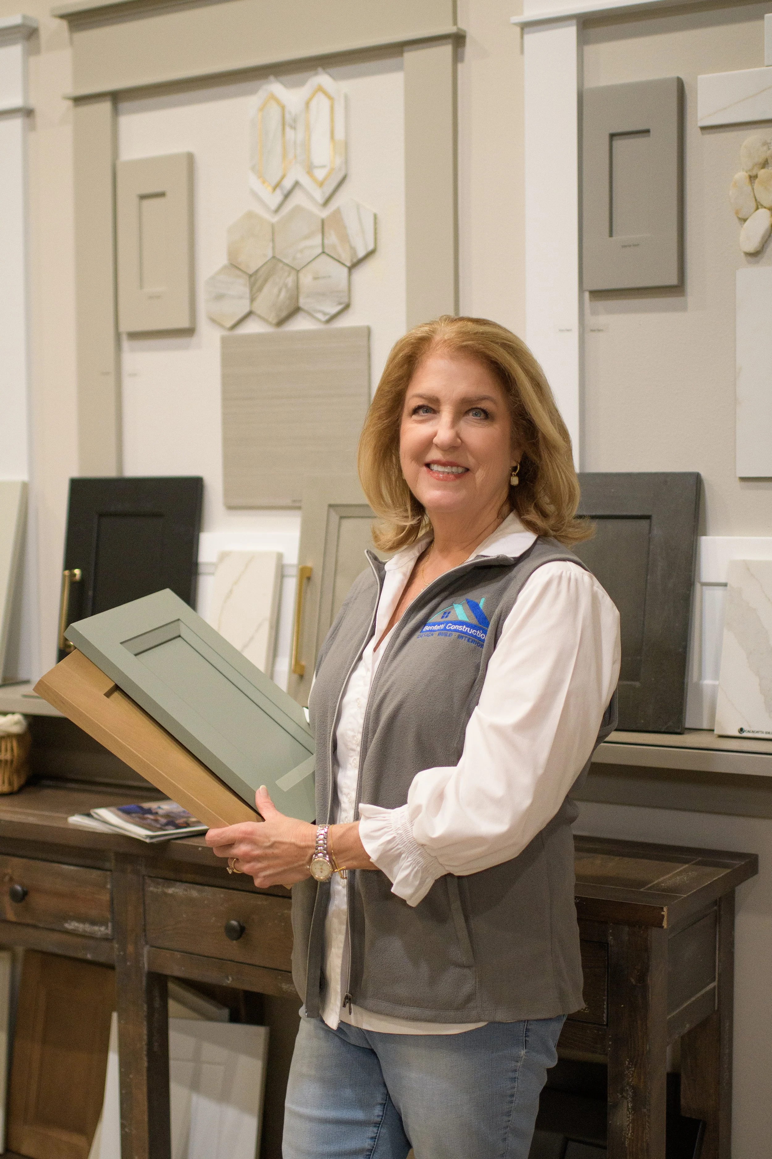 A woman standing in a showroom holding a cabinet door sample, surrounded by various cabinet doors and samples on the wall behind her.