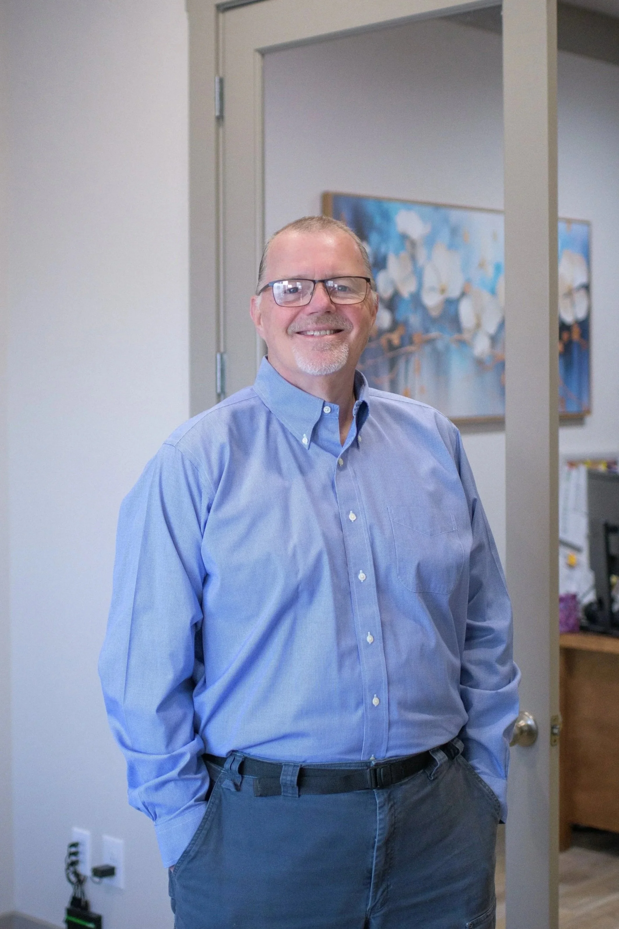A smiling man with glasses and a light beard, wearing a blue button-up shirt and dark pants, stands in an office with a closed door behind him and a painting on the wall.