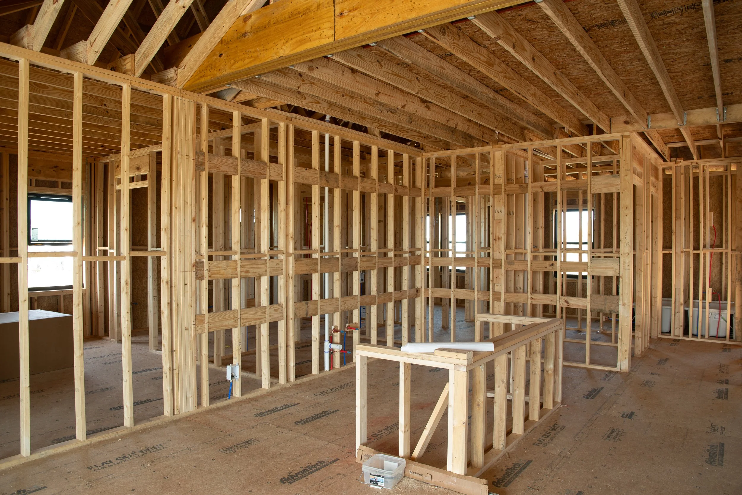 Interior view of a house under construction showing wooden framing, exposed ceiling beams, and construction materials.