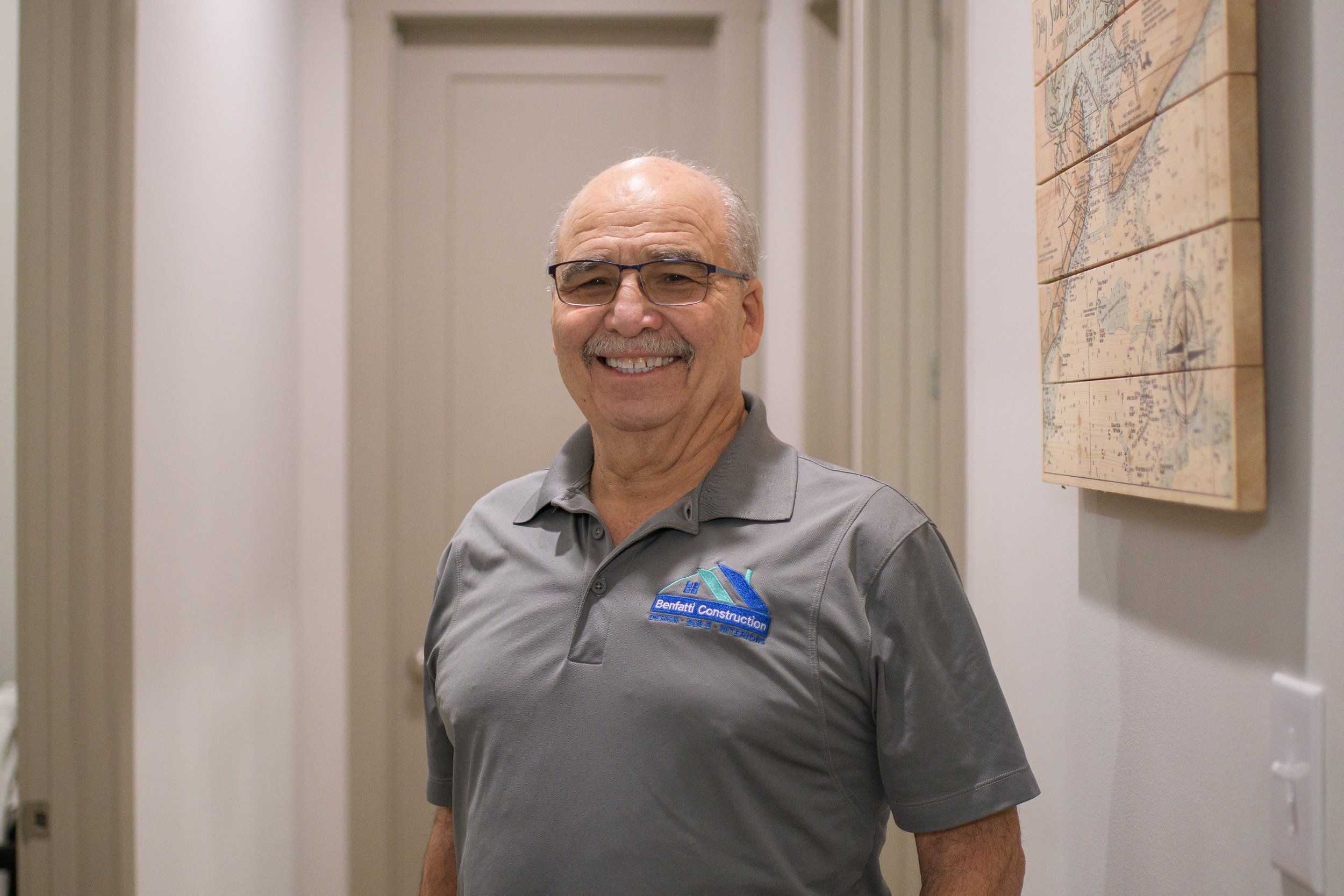 A smiling older man with glasses and a mustache wearing a gray polo shirt with a company logo that says "Benfatti Construction" standing indoors next to a wall with a map or decorative picture.