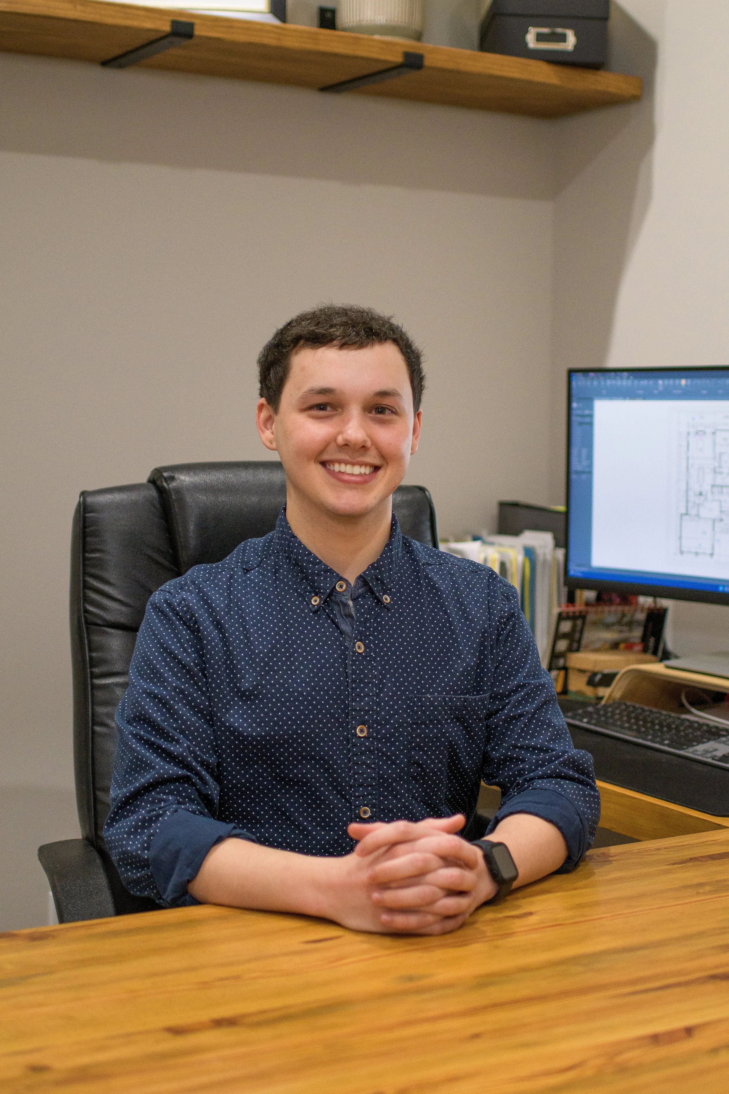 A young man with short dark hair smiling, wearing a navy blue polka dot button-up shirt and a smartwatch, sitting at a wooden desk in an office, with a computer monitor and office supplies in the background.