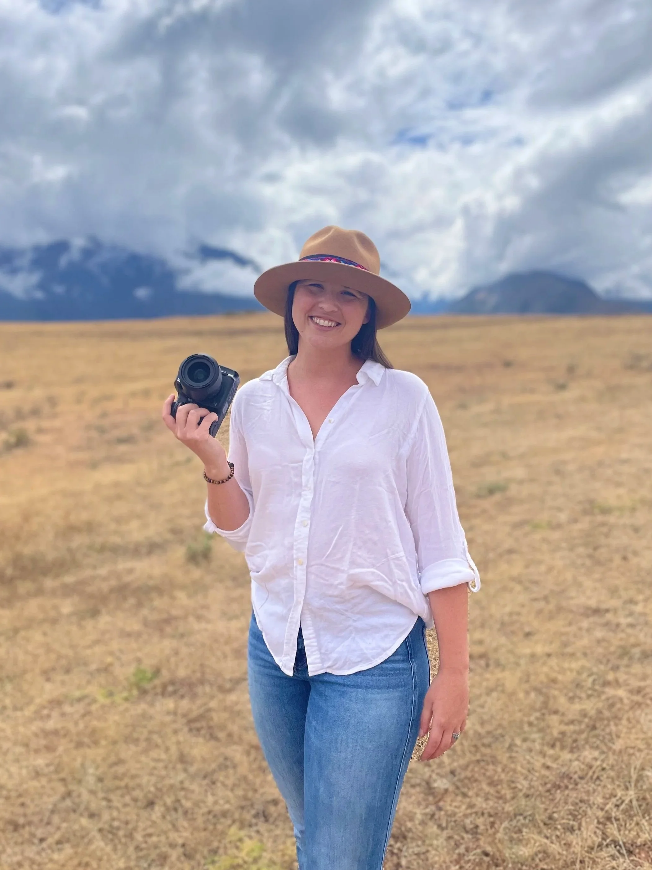 Woman standing outdoors in a field, holding a camera, wearing a wide-brimmed hat and a white shirt with rolled-up sleeves, smiling at the camera with mountains and cloudy sky in the background.
