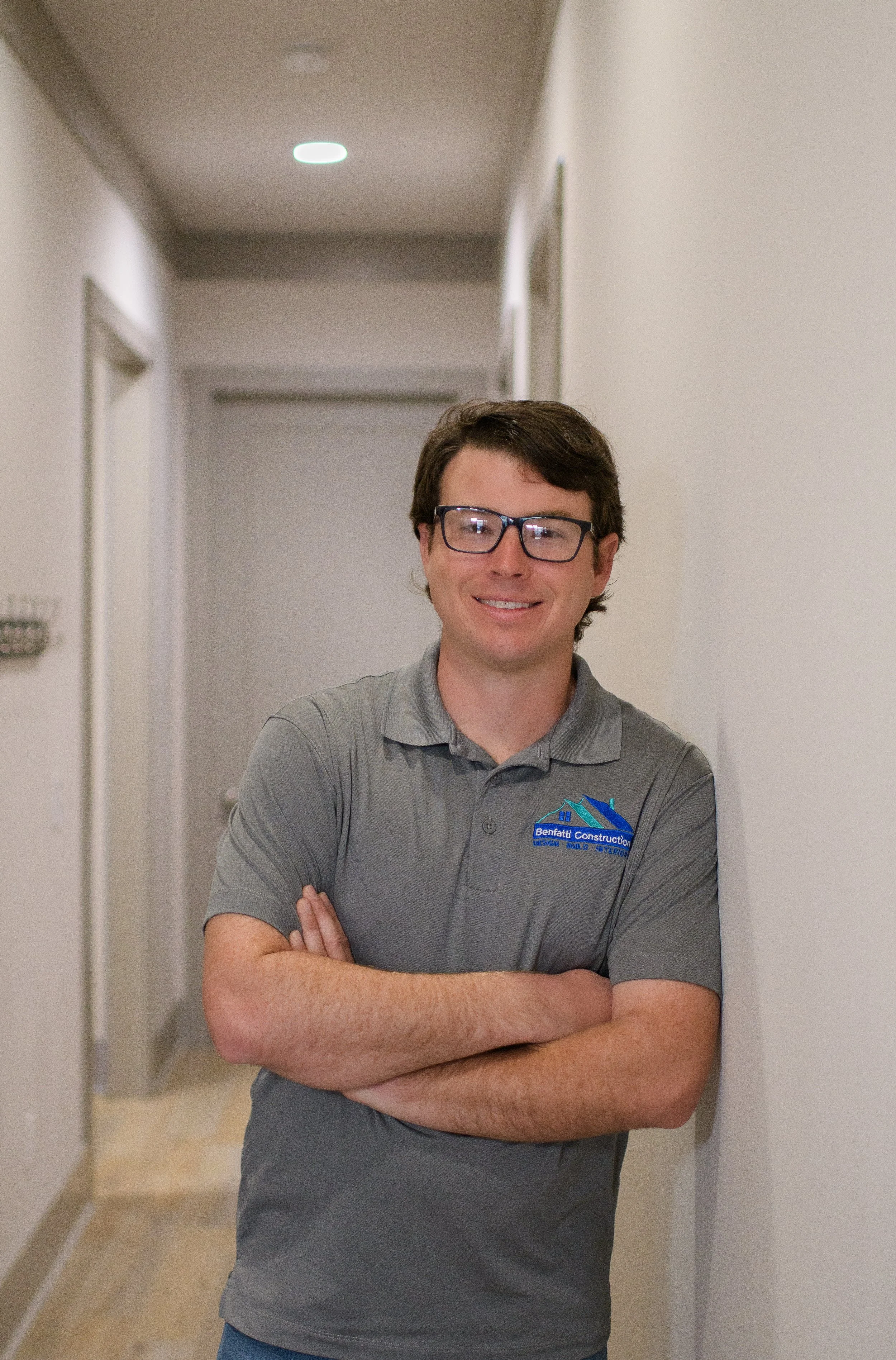 A man wearing glasses and a gray polo shirt with a construction company logo, smiling and standing with arms crossed in a hallway.