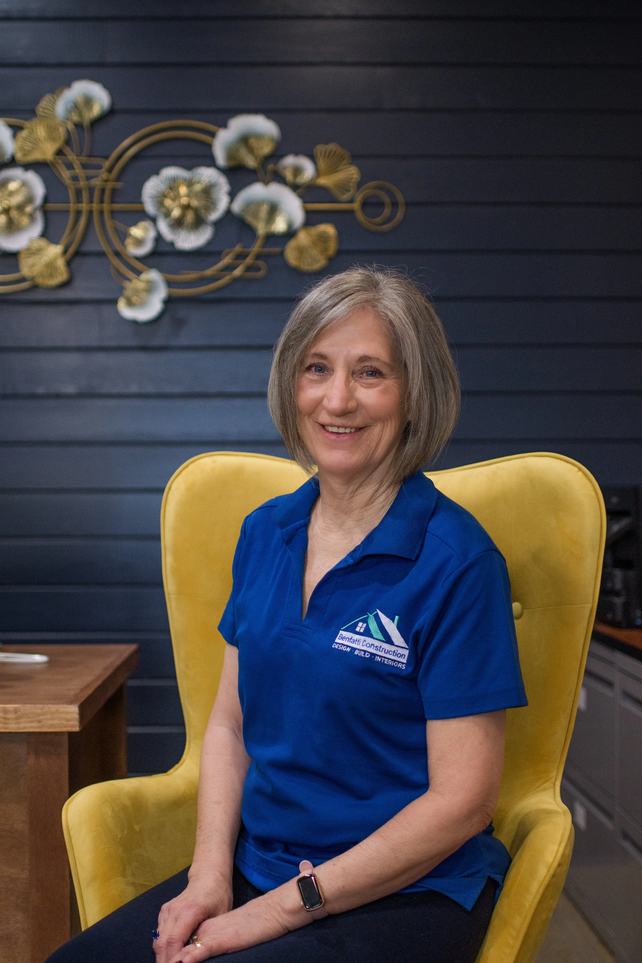 A smiling middle-aged woman with short gray hair sitting in a yellow armchair, wearing a blue polo shirt with a construction company logo, in a room with dark blue paneled walls and decorative wall art.