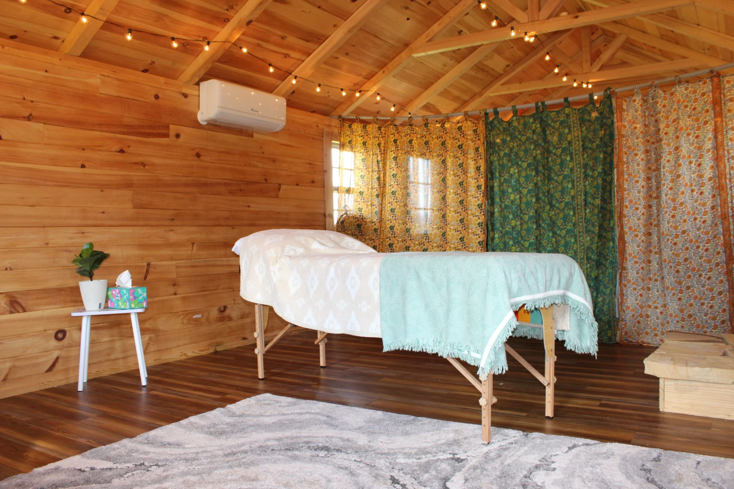 A cozy massage or reiki room with wooden walls and ceiling, string lights, an air conditioner, a massage table with white and light blue blankets, a small side table with a plant and tissues, colorful curtains, and a patterned rug.
