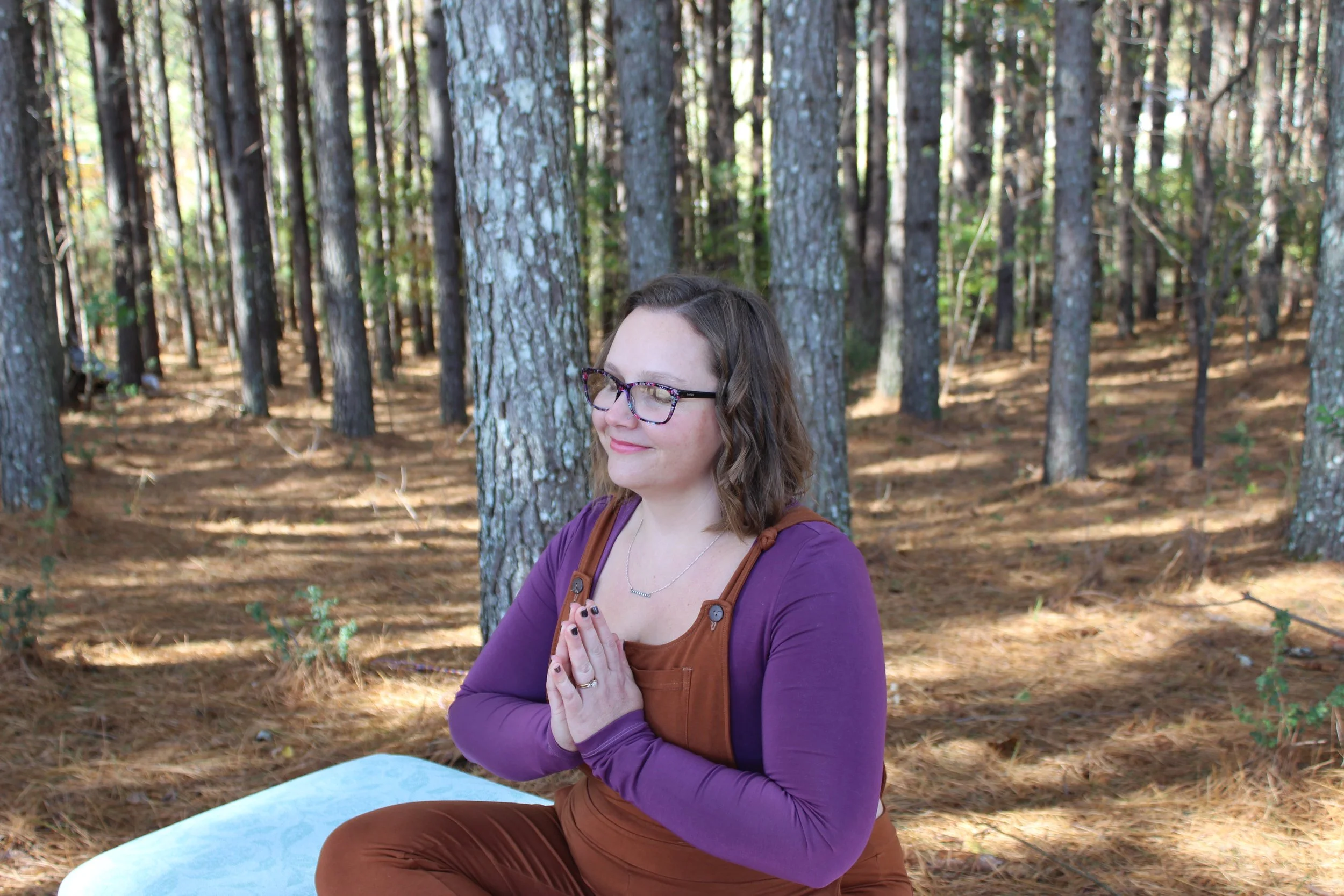A woman with glasses and short brown hair practicing yoga or meditation outdoors in a forest, sitting cross-legged with her hands in a prayer position, eyes closed, and a slight smile.
