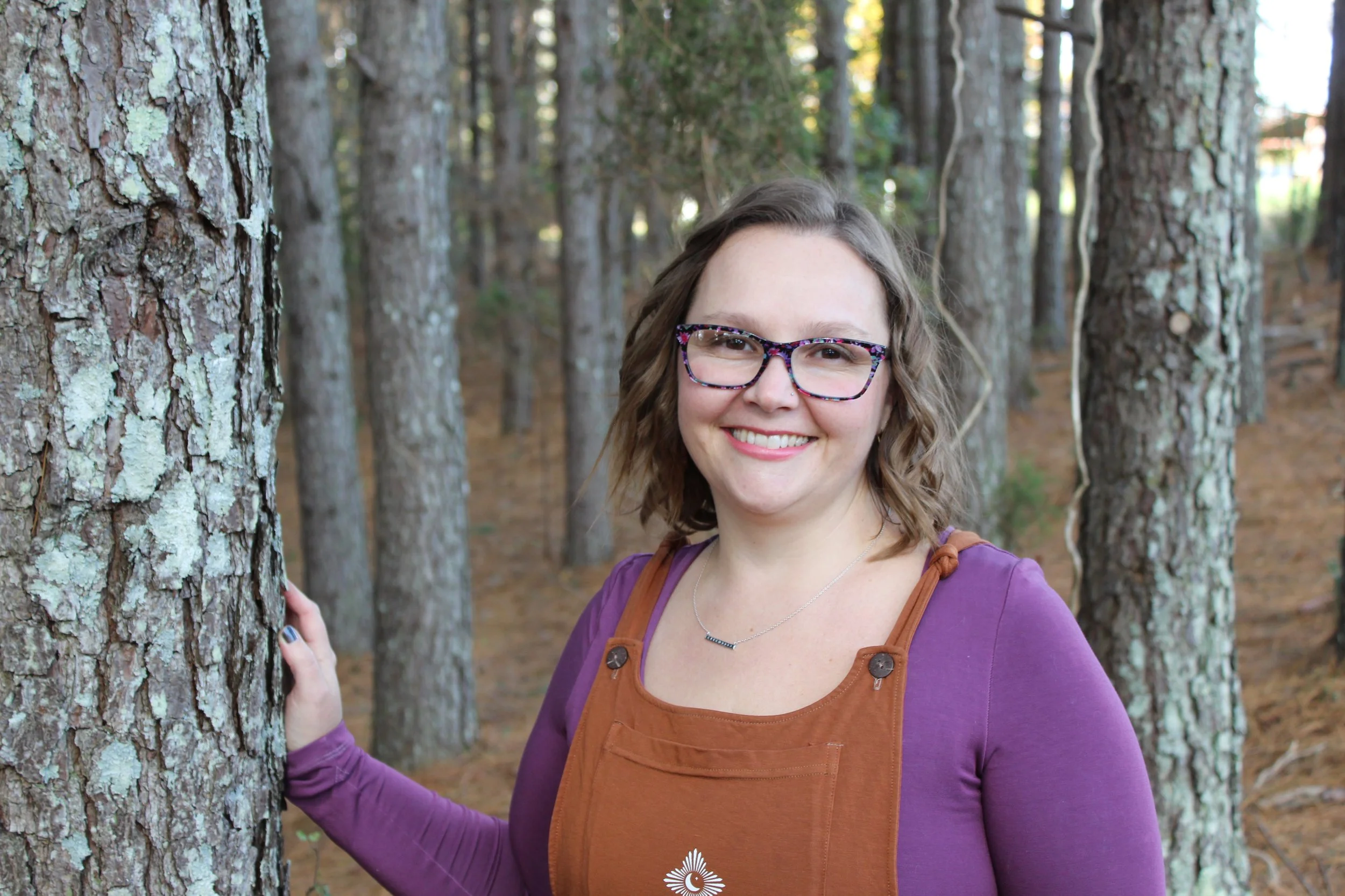 A woman with glasses and wavy brown hair smiling outdoors among trees, wearing a purple long sleeve shirt and a brown overalls.
