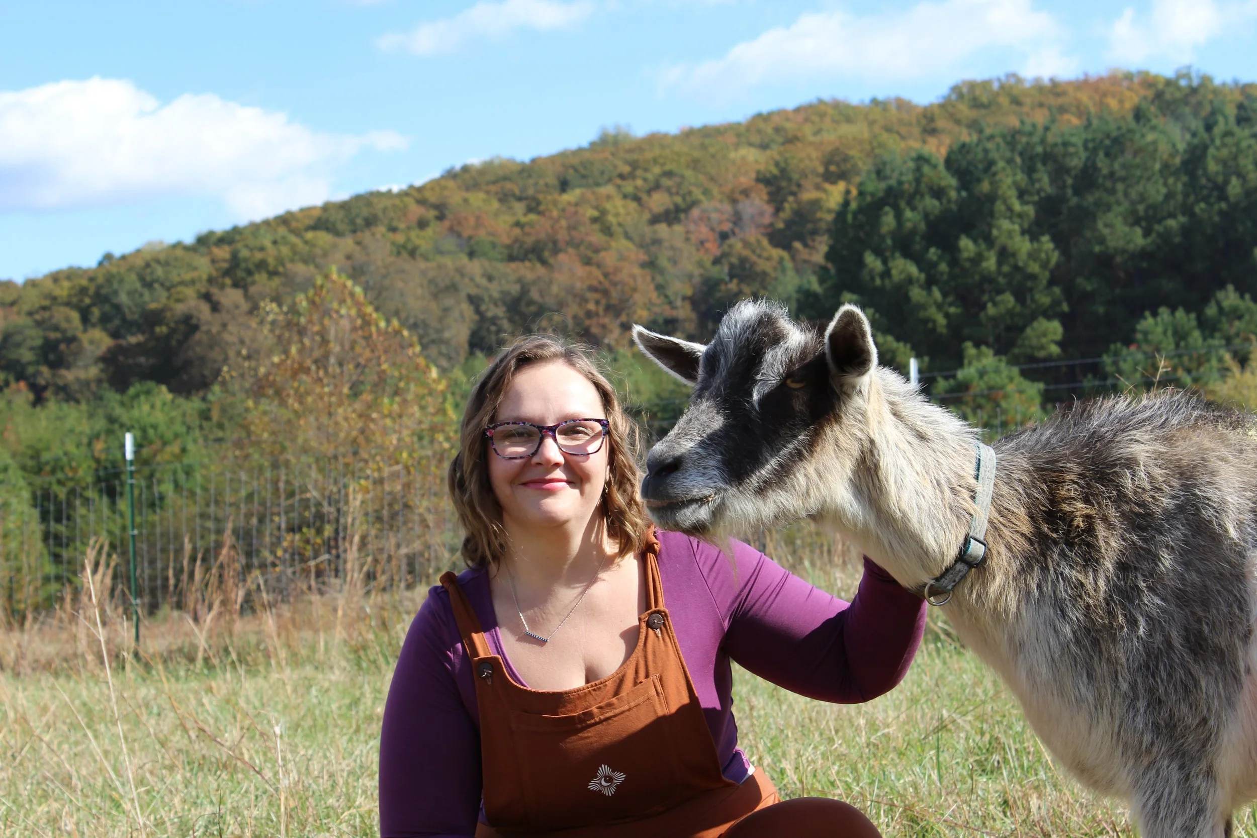 A woman with glasses smiling while petting a goat in a grassy field with trees and hills in the background on a sunny day.