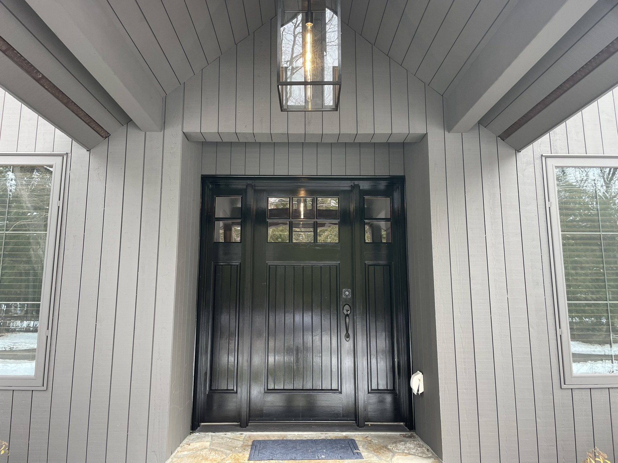 Black front door with windows at the top, flanked by two windows on a gray house exterior with vertical siding. A rectangular light fixture hangs above the door.